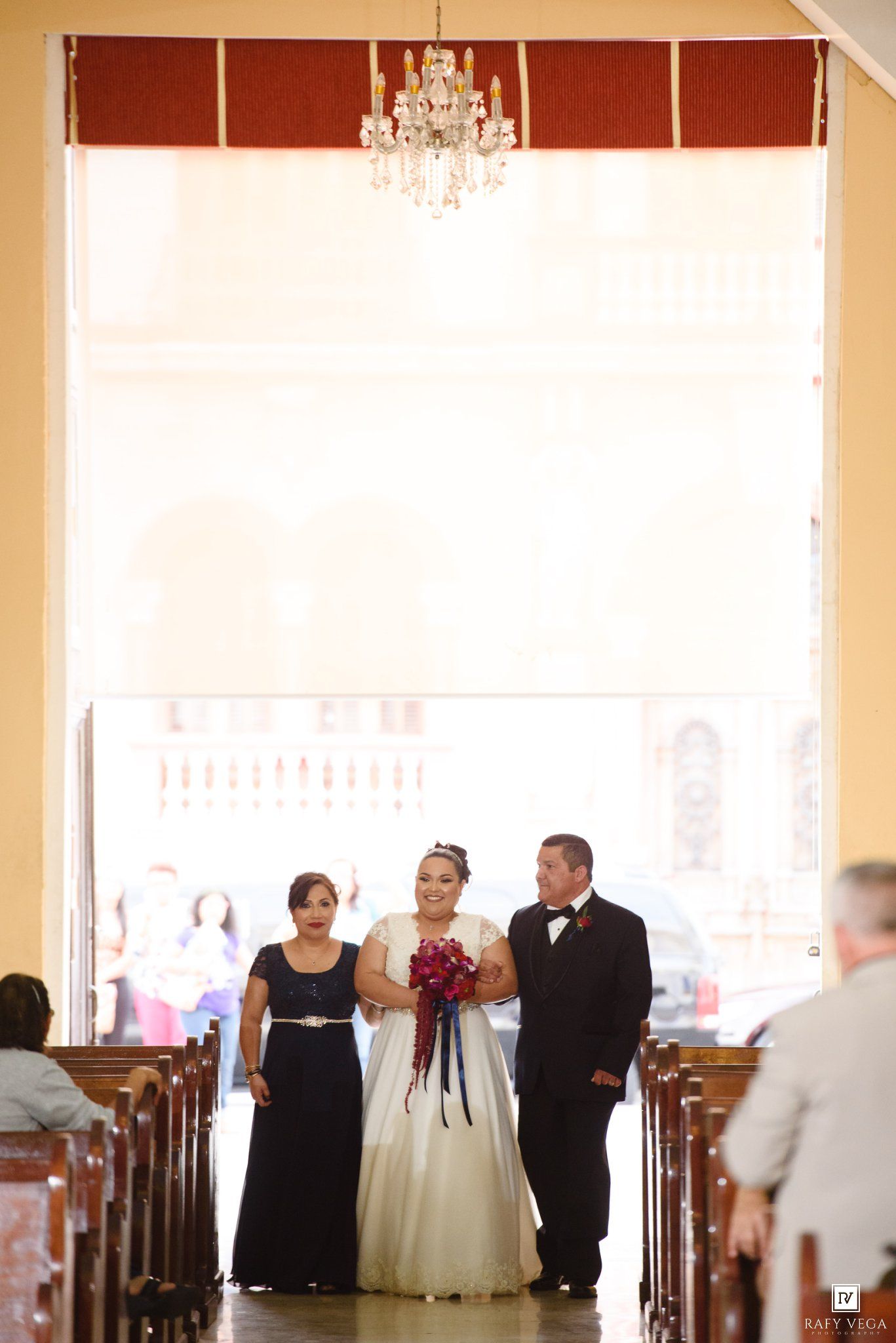 Bride in white gown walks down church aisle with parents, holding red bouquet, chandelier above.