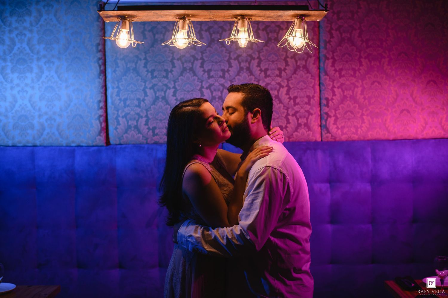 A couple shares a romantic moment, nearly kissing under warm lights in a vibrant blue and pink lit lounge.
