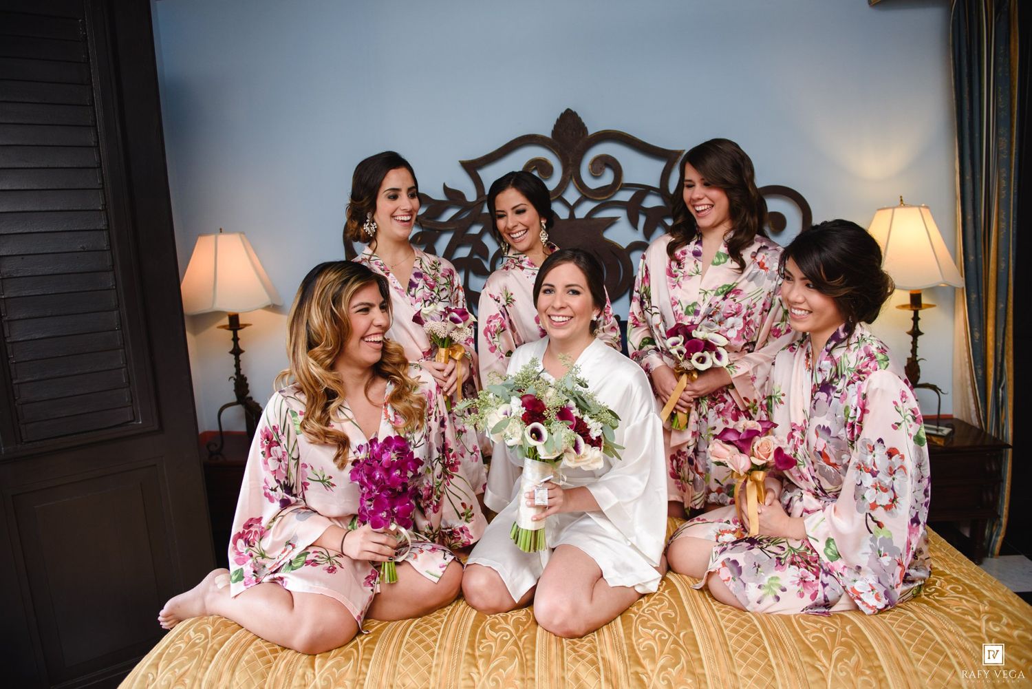 Bride and bridesmaids in floral robes holding bouquets, smiling together on a bed before the wedding ceremony.