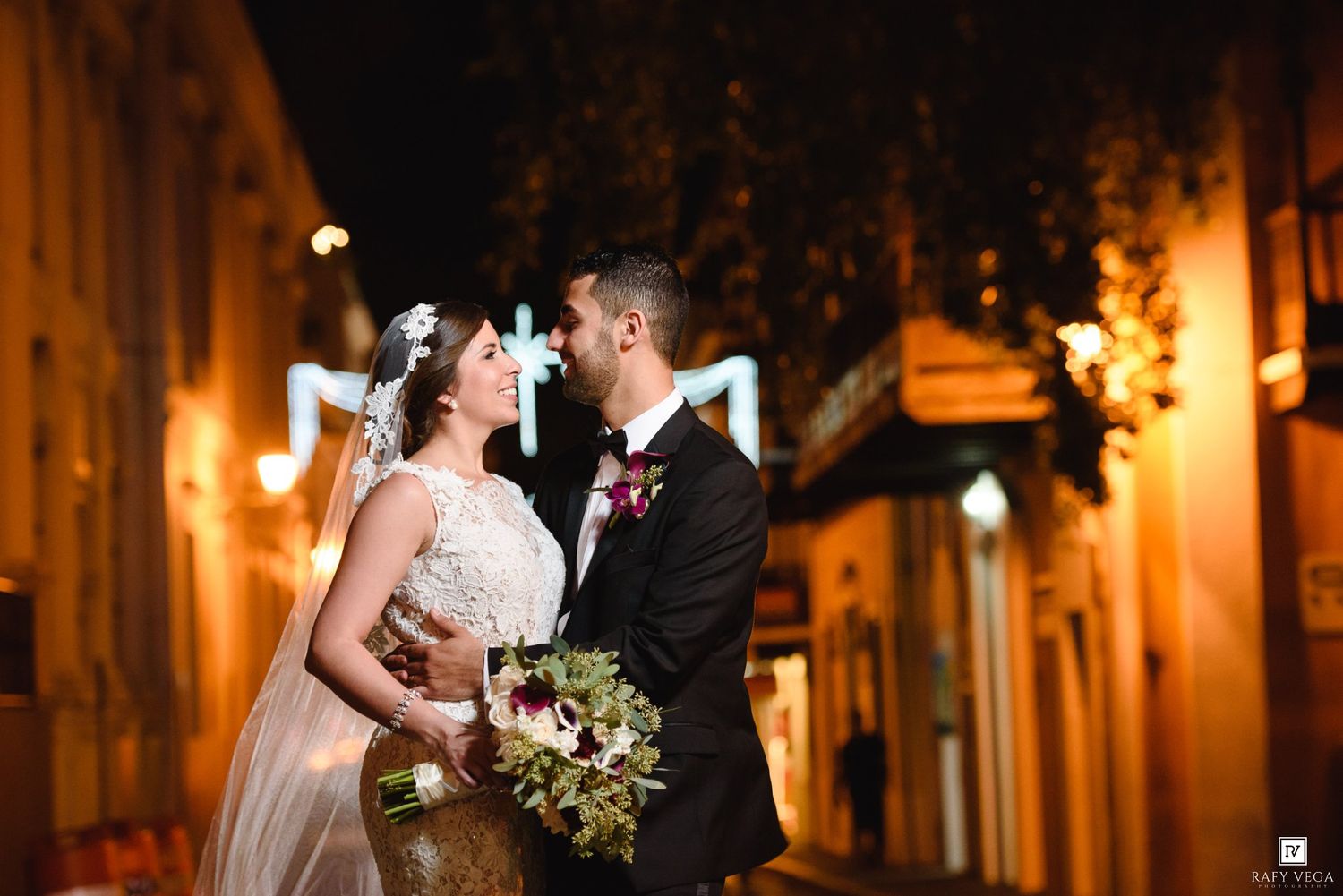 Bride and groom share a romantic moment on a warmly lit street at night, with the groom in a suit and bride in a lace gown.