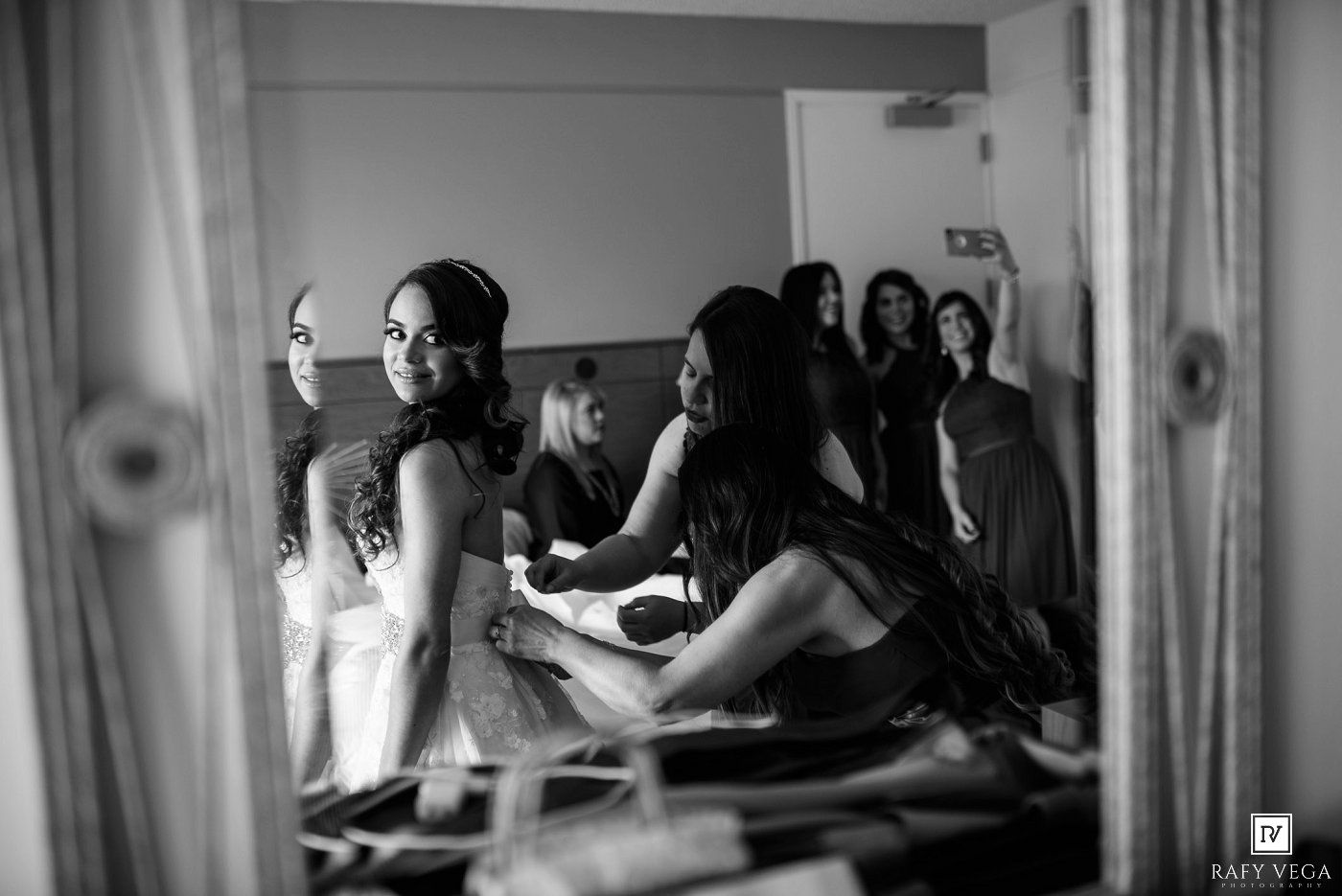Black and white photo of bride getting ready with bridesmaids helping in a mirror-reflected room.