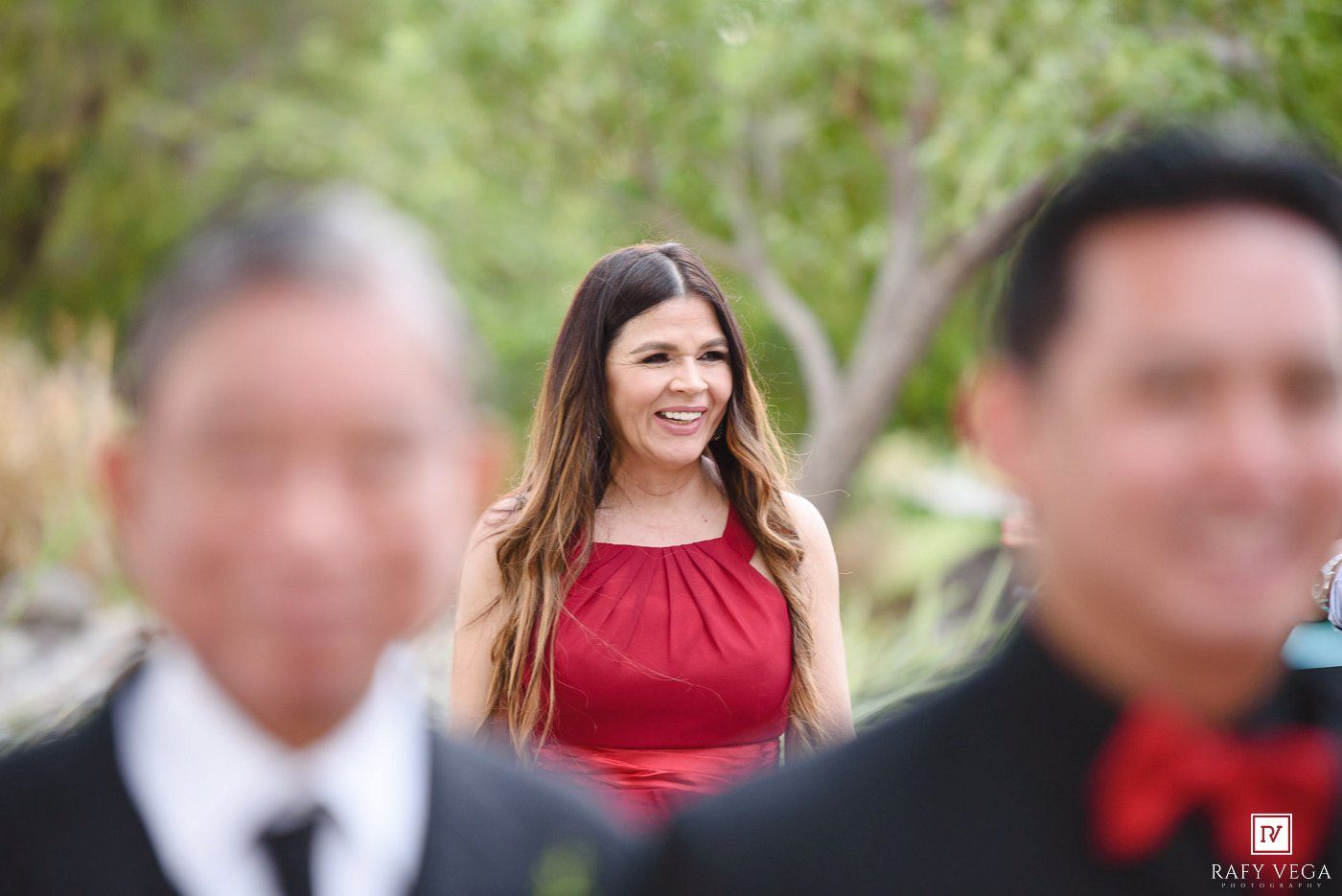 Smiling woman in red dress stands between two blurred men in suits at wedding.