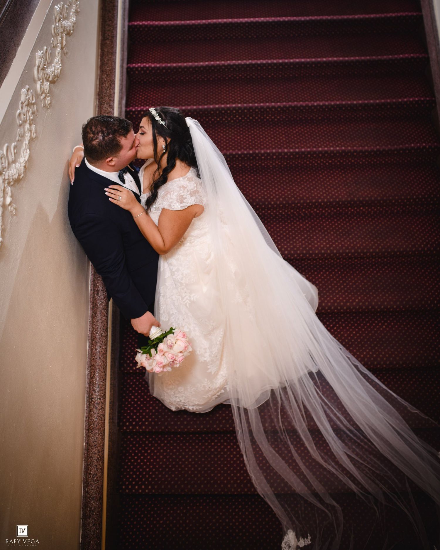 Bride and groom share a romantic kiss on a grand staircase, bride in lace gown with veil holding pink bouquet.