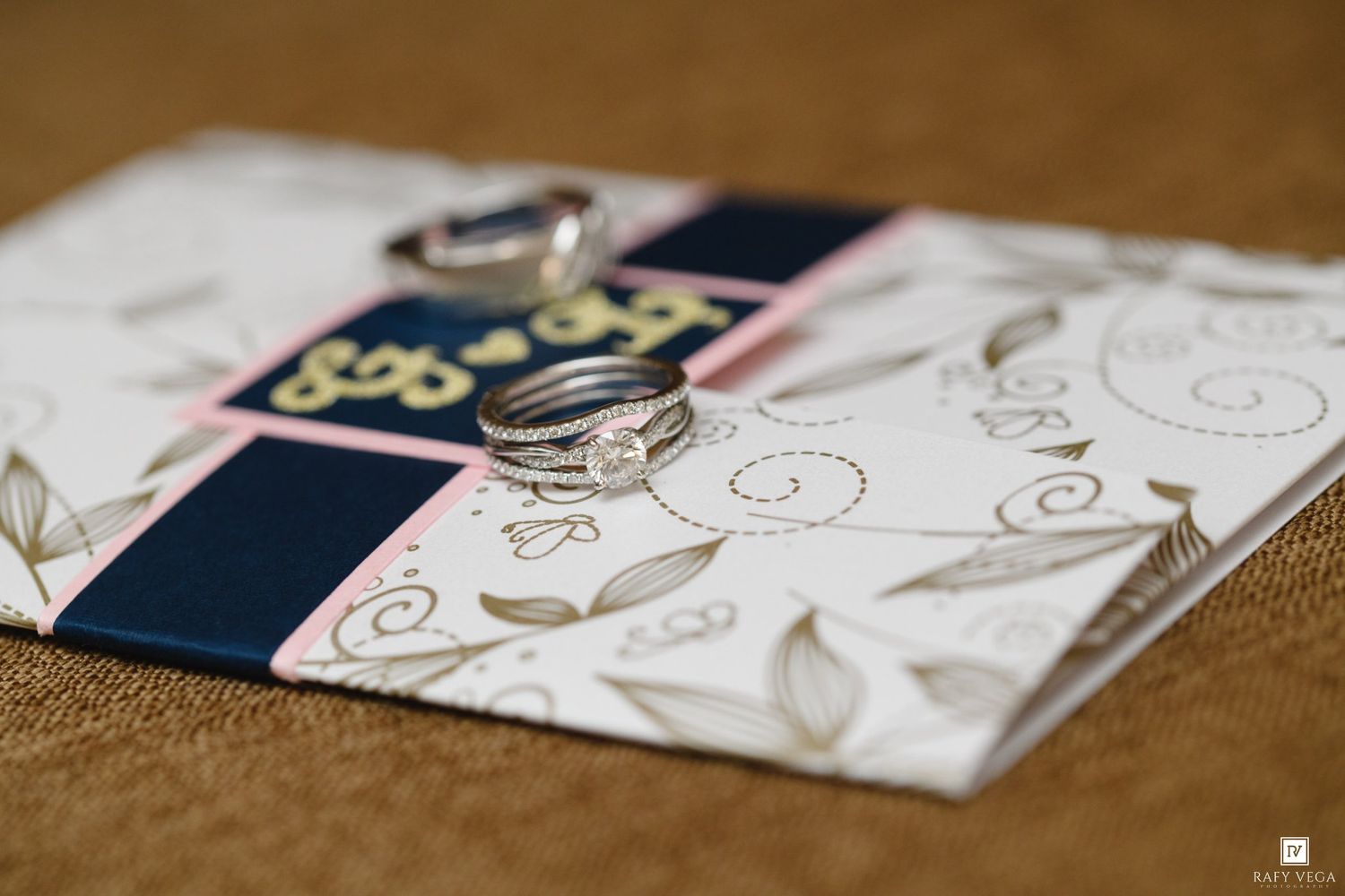 Two wedding rings resting on a floral white, navy, and pink wedding invitation card on a burlap surface.