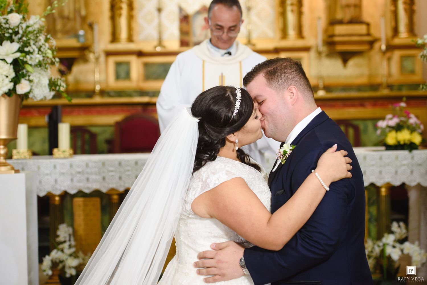 Bride and groom share their first kiss at the altar during a church wedding ceremony with priest officiating.