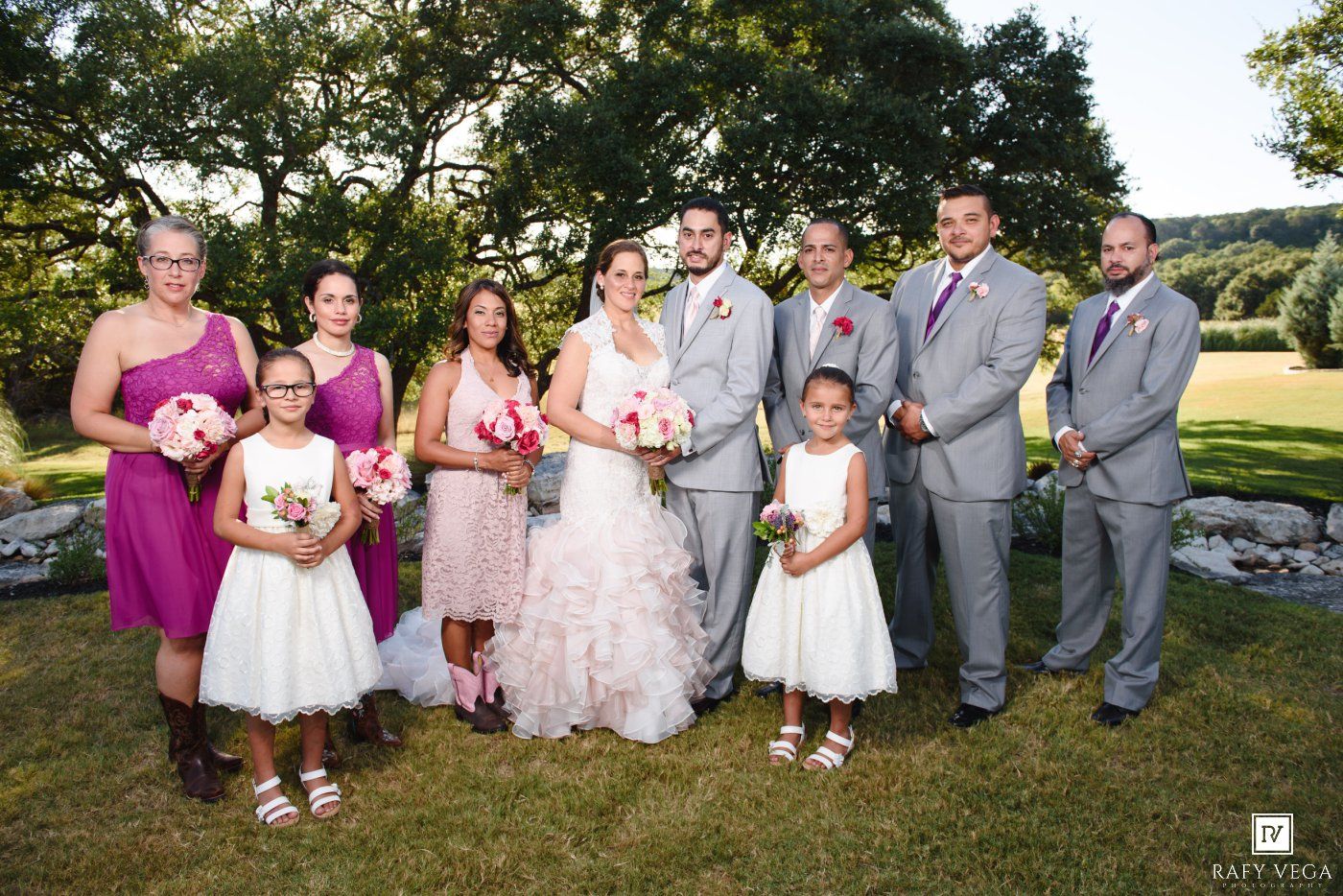 Wedding party poses outdoors on green lawn, bride in white gown with bridesmaids in purple dresses and groomsmen in gray suits.