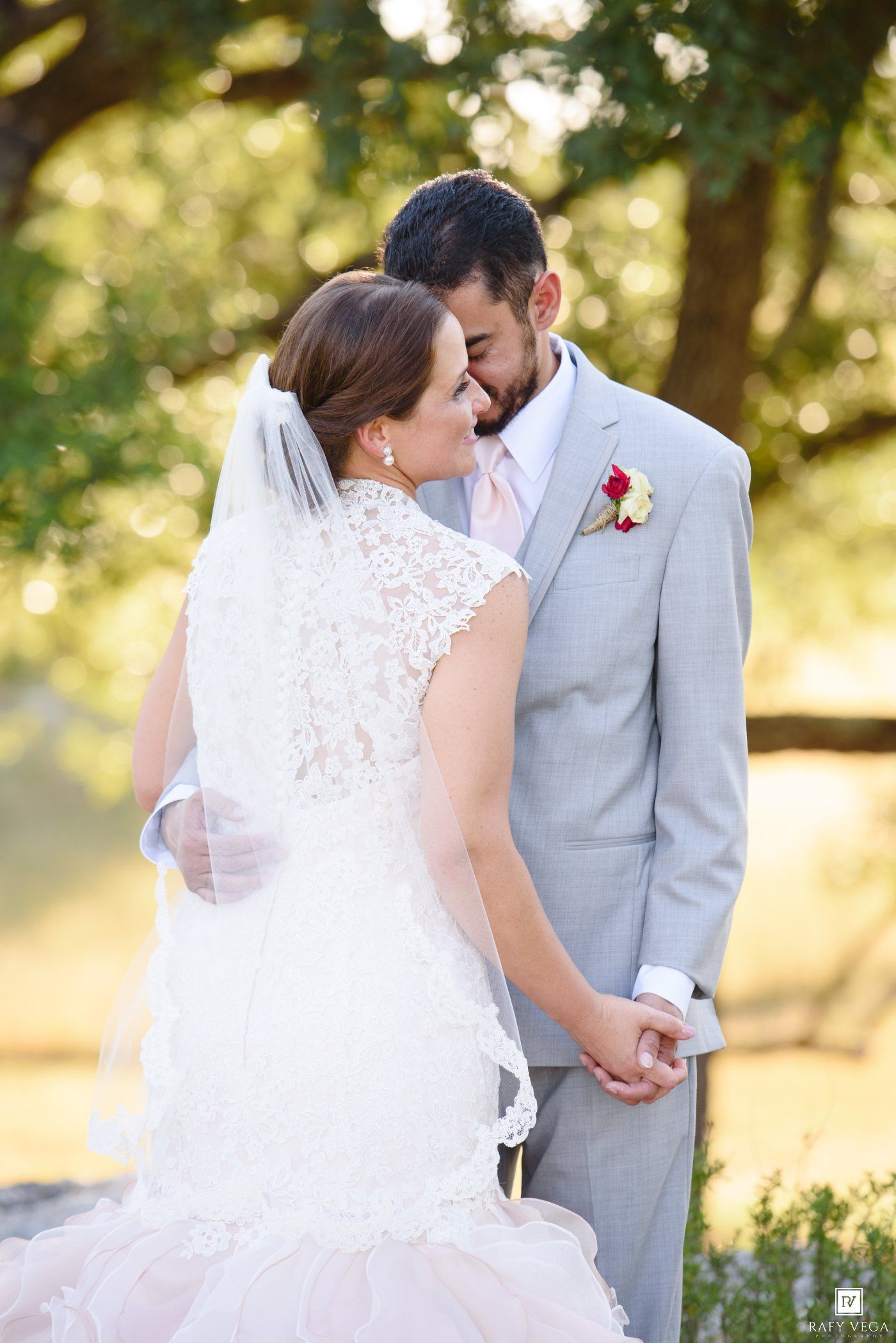 Bride and groom share a romantic moment outdoors, bride in lace veil and gown, groom in gray suit with pink boutonniere.