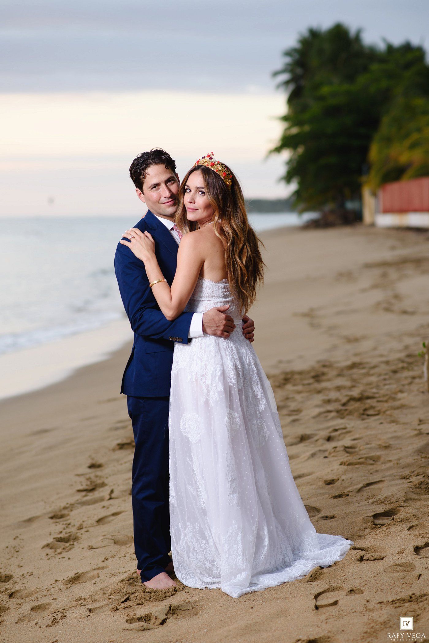 Couple in wedding attire embracing on a tropical beach at sunset, bride in white gown with floral crown.