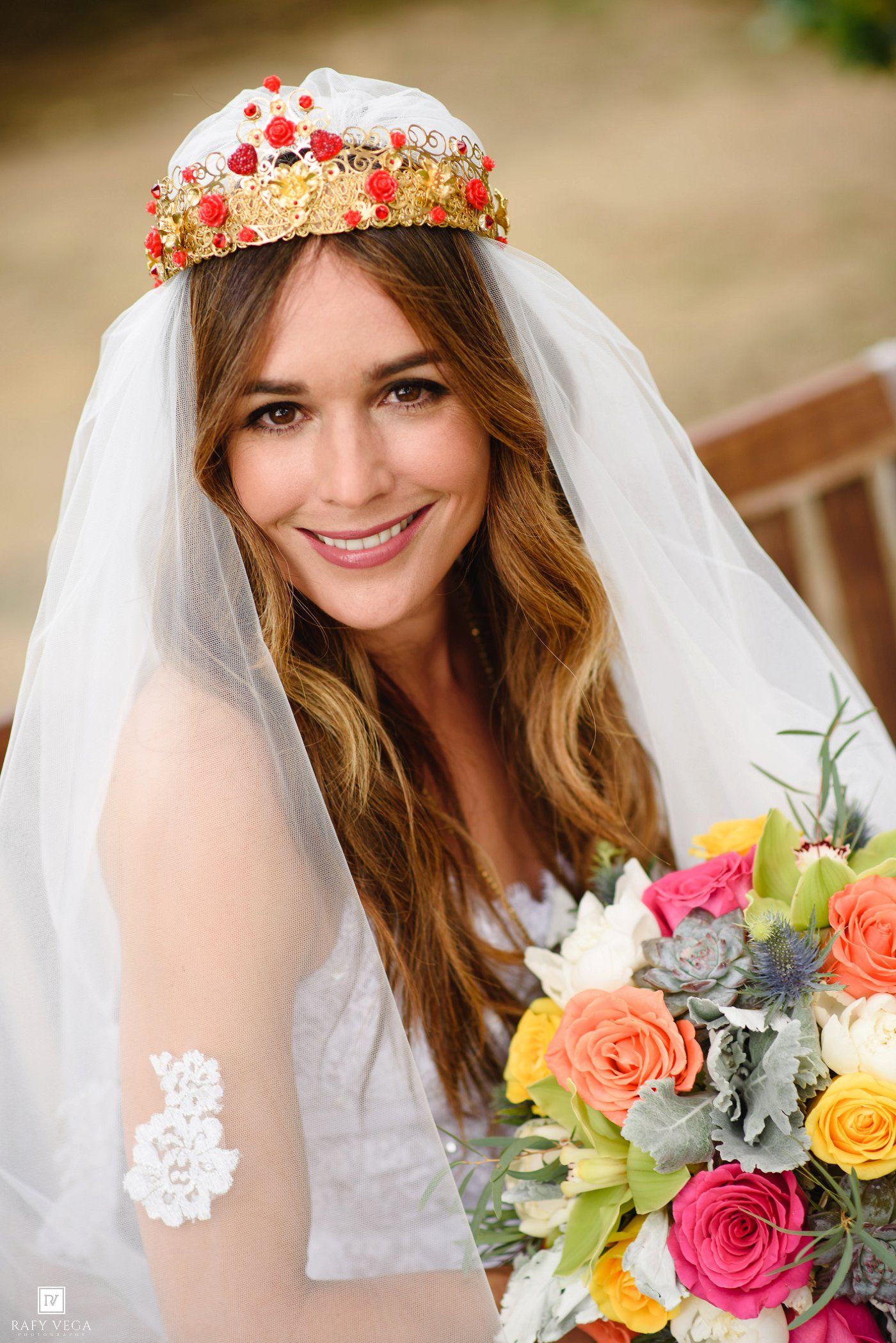 Smiling bride wearing a floral crown and white veil holds a colorful bouquet of roses and succulents.