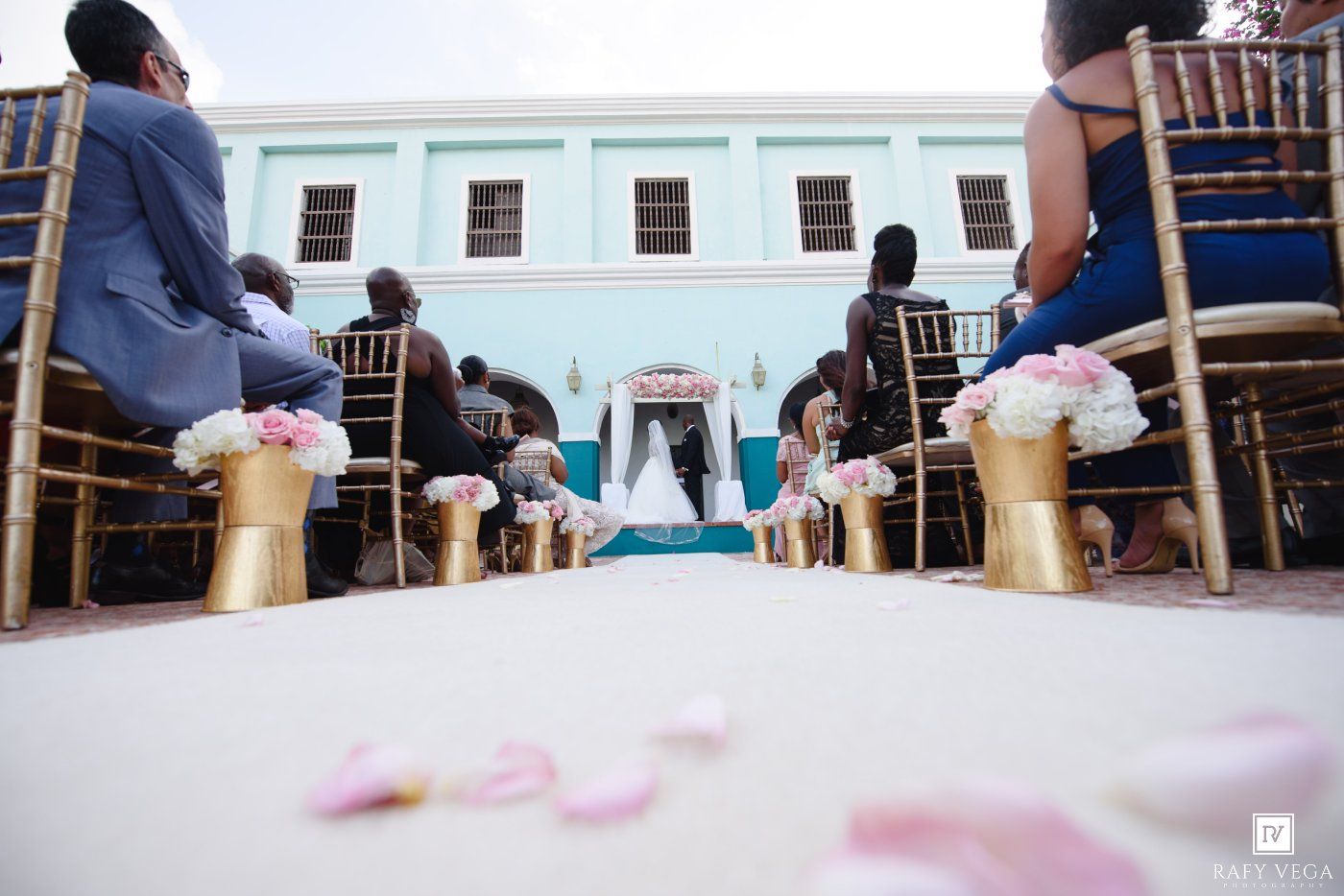 Outdoor wedding ceremony aisle with gold vases, pink flowers, rose petals, and guests seated on blue chairs before an altar.