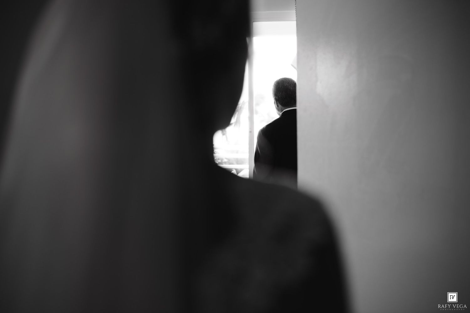Black and white wedding photo of groom silhouetted in doorway, captured through narrow gap between walls.