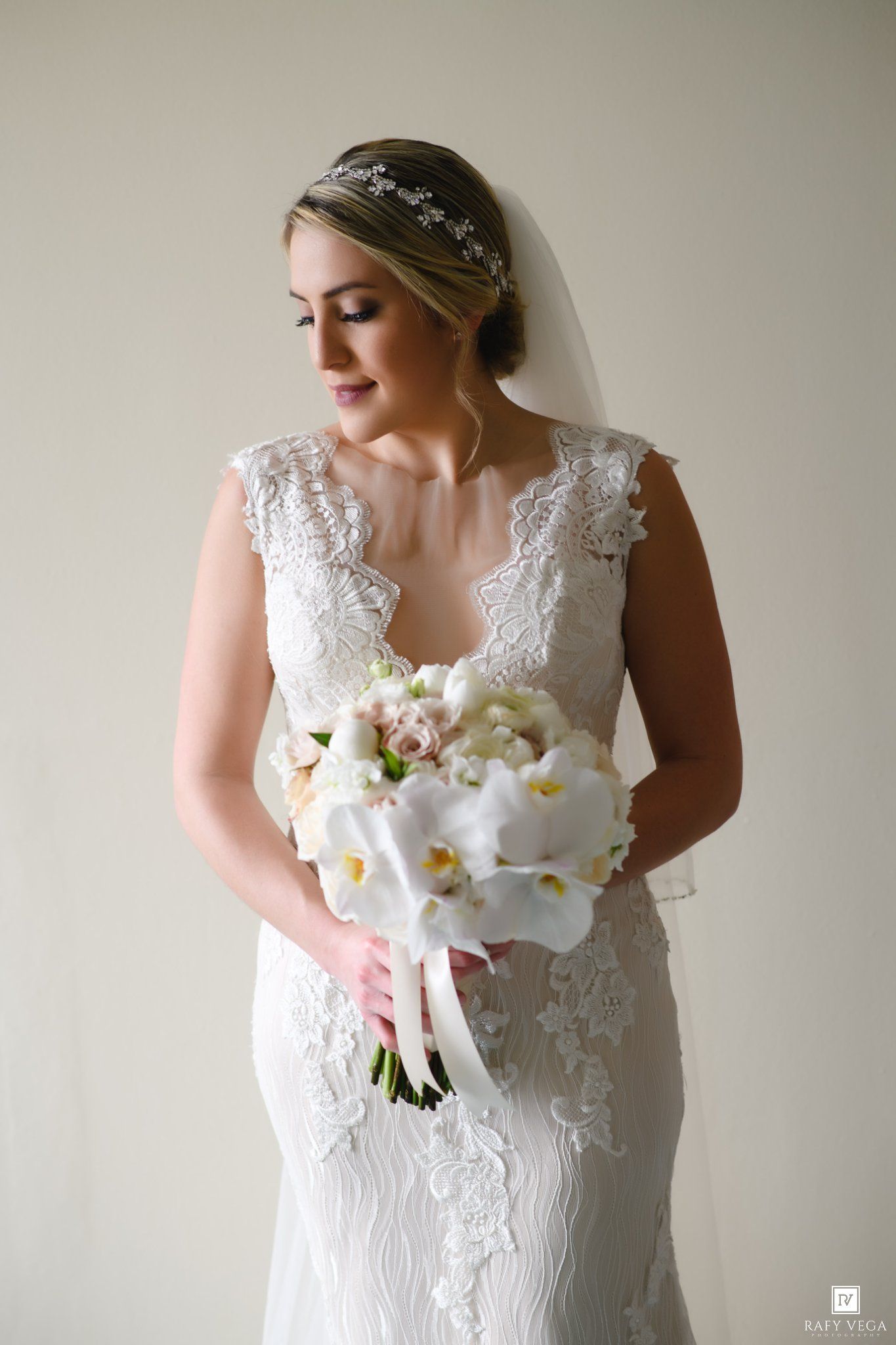Bride in lace wedding gown holding white orchid bouquet with ribbon, wearing veil and floral hairpiece.
