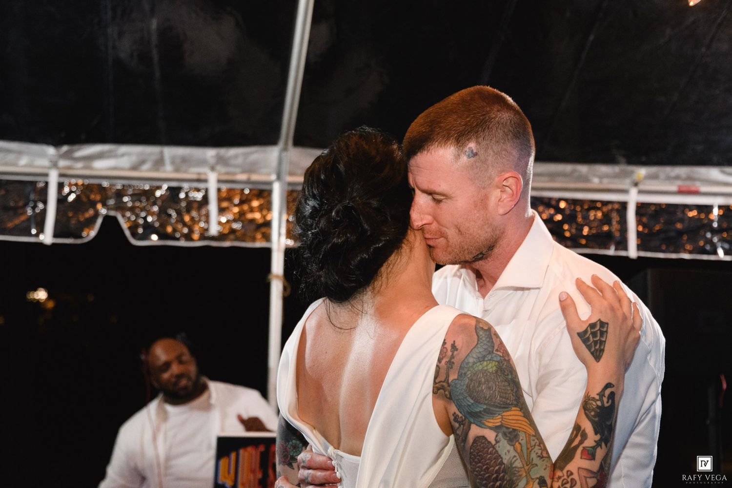 Bride and groom share first dance under tent at outdoor wedding reception, groom with tattoos in white shirt.