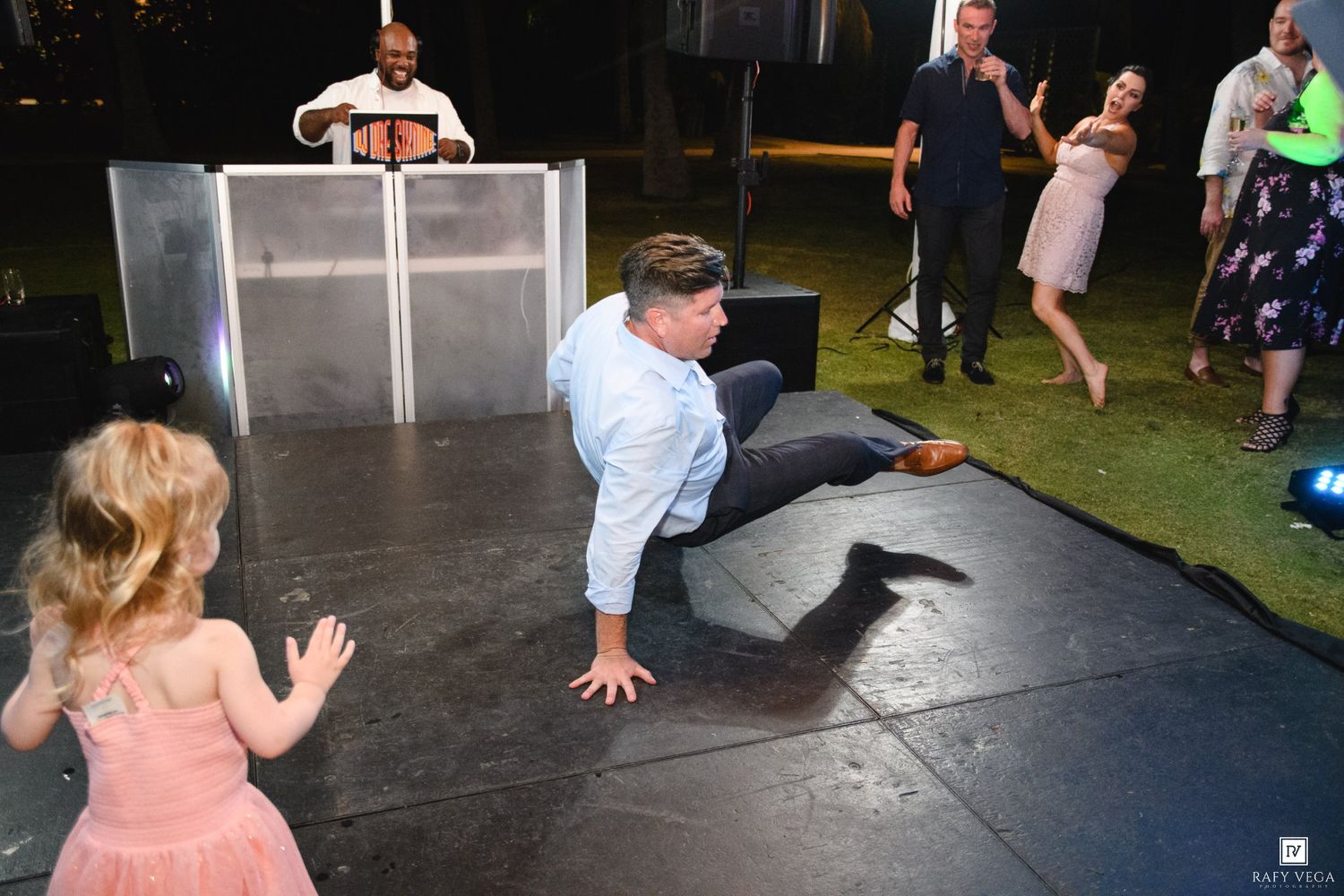 Man breakdancing at wedding reception while child watches and DJ performs in background.