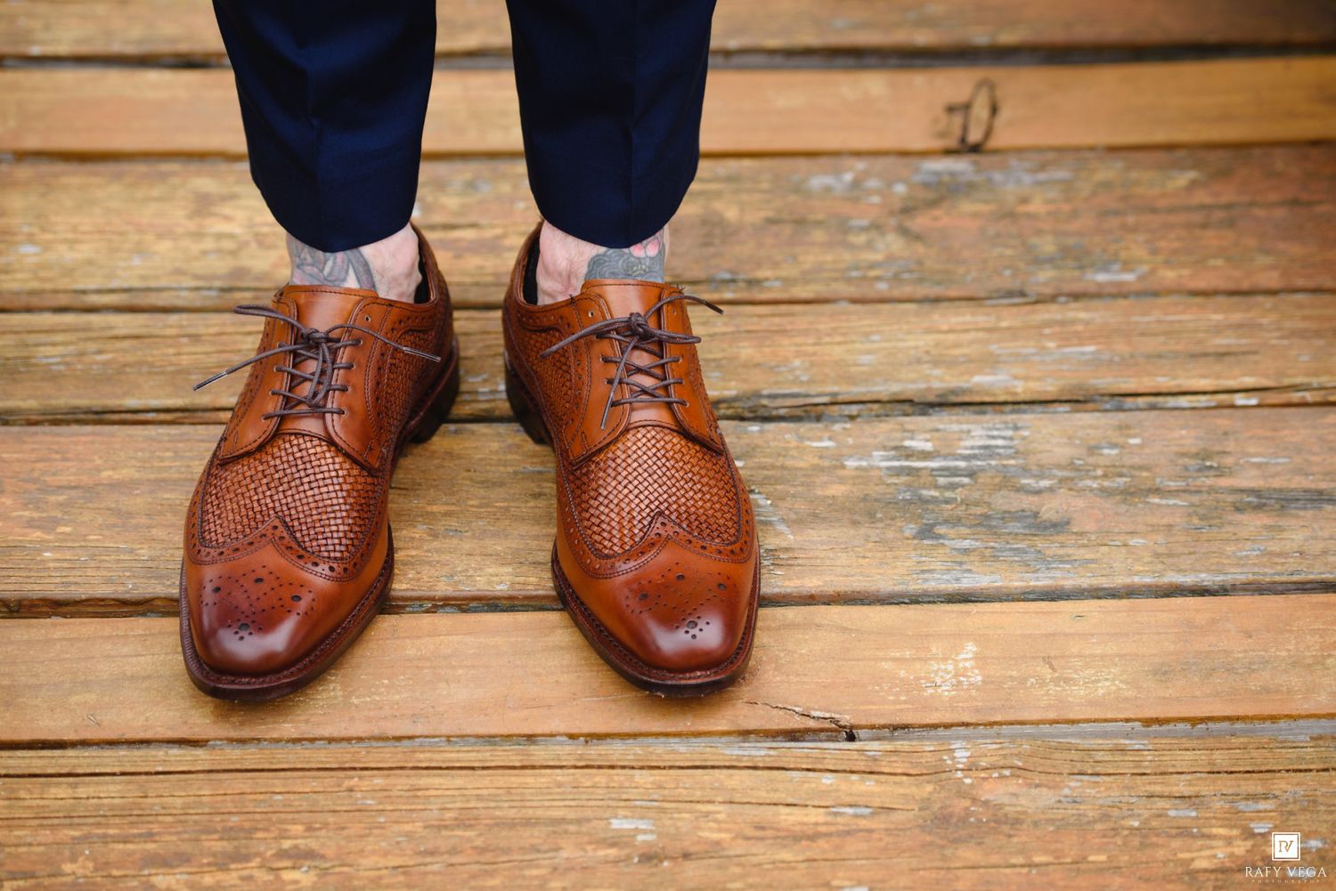 Close-up of men's tan leather brogue Oxford dress shoes on rustic wooden floor with navy trousers.