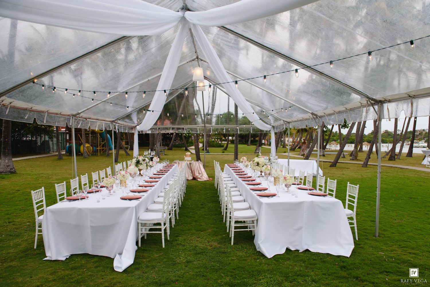 Elegant outdoor wedding reception under a white tent with long tables, white chairs, and string lights on a green lawn.