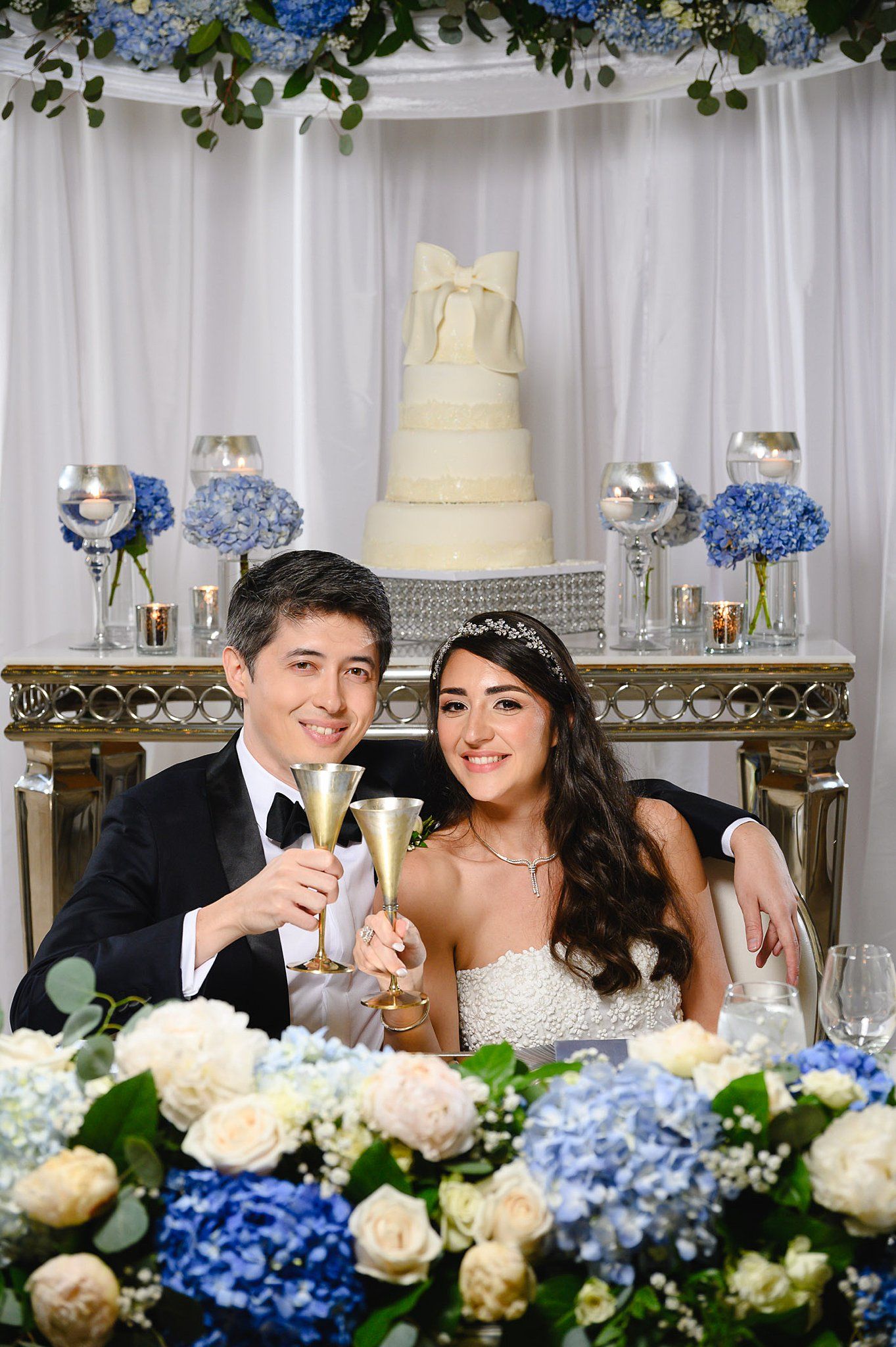 Bride and groom toast with champagne flutes at sweetheart table adorned with blue hydrangeas and white wedding cake.