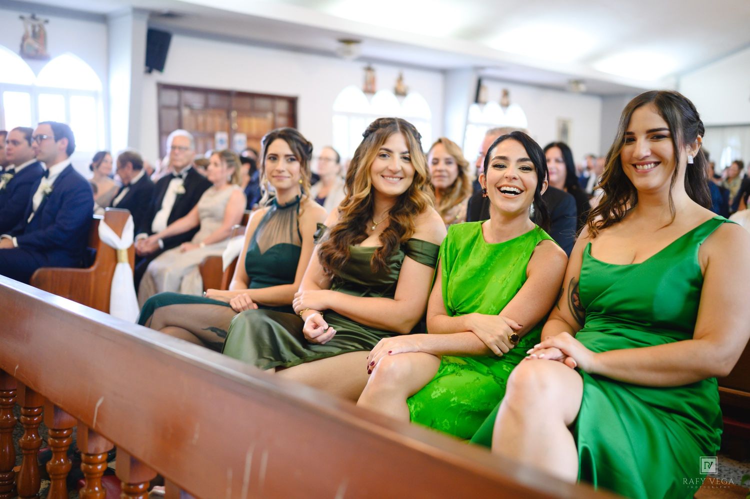 Women in green dresses laughing and smiling together in church pews during a wedding ceremony.