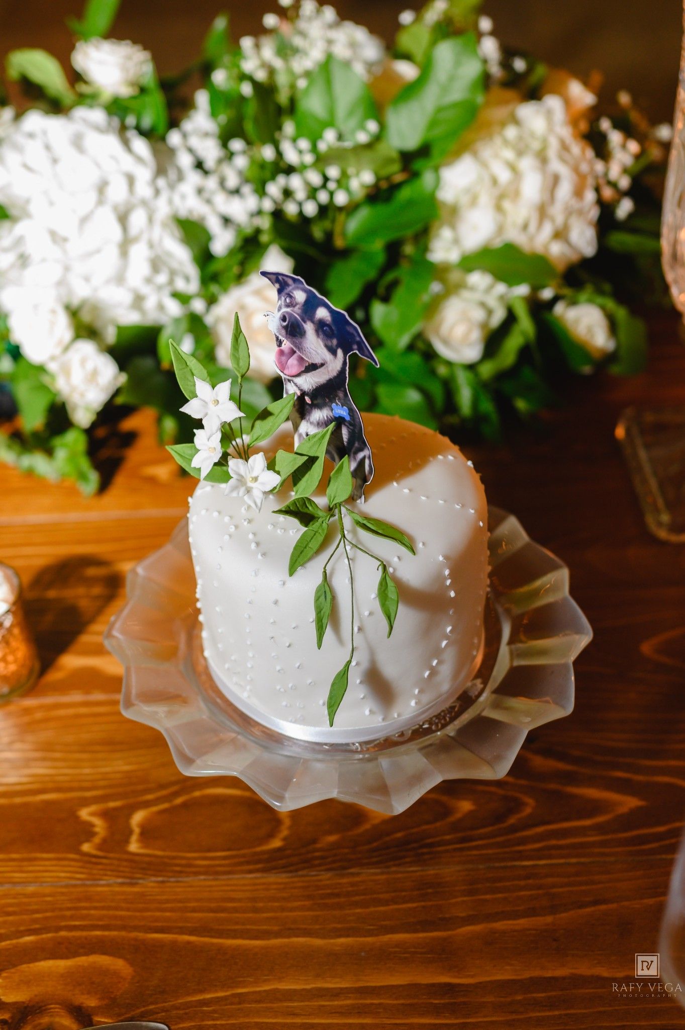 White fondant wedding cake with dog topper and floral decorations on wooden table.
