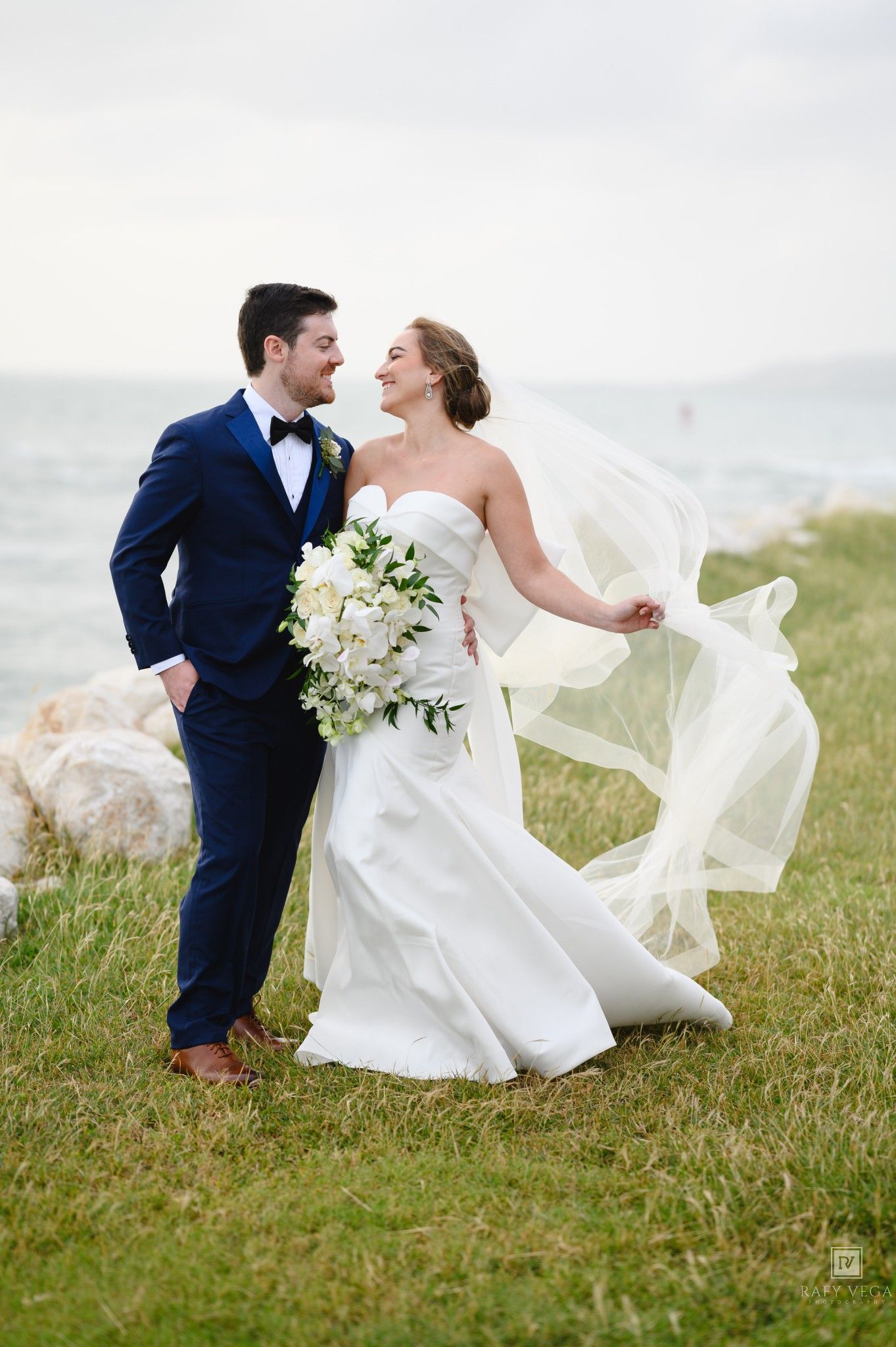 Bride and groom share a romantic moment outdoors by the water, her veil flowing in the wind, as she holds a white floral bouquet.