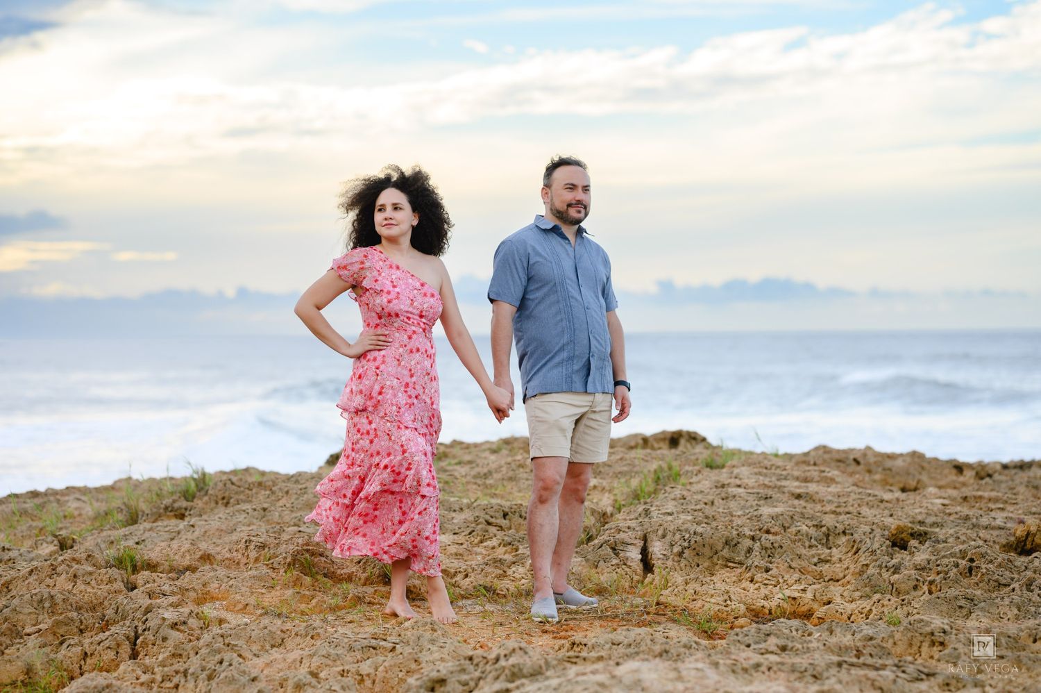 Couple holding hands on rocky seaside cliffs, woman in floral dress, man in blue shirt.