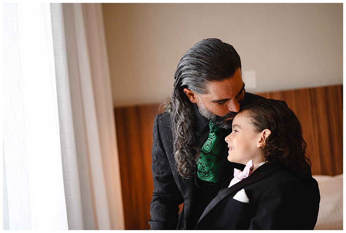 Groom and young boy in matching black tuxedos share a tender moment by a window before a wedding.