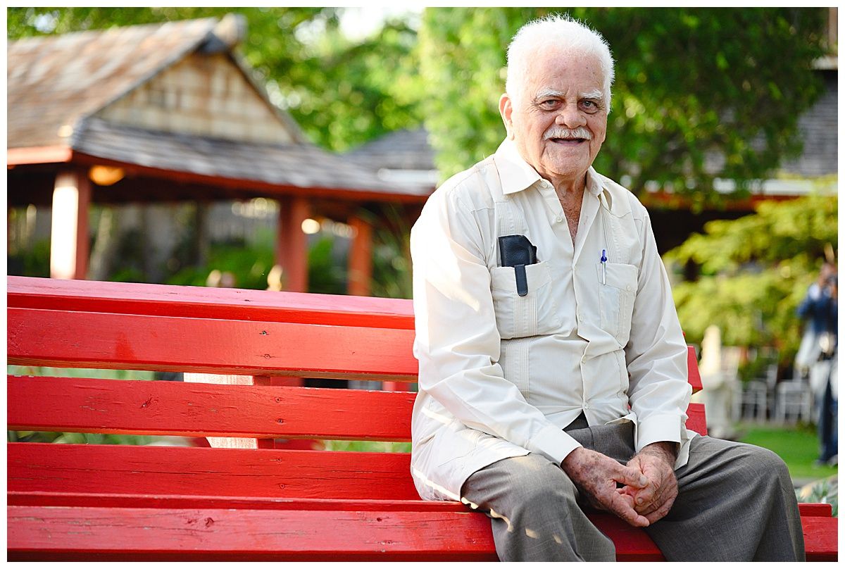 Elderly man with white hair sitting on a red wooden park bench, smiling outdoors among green trees.