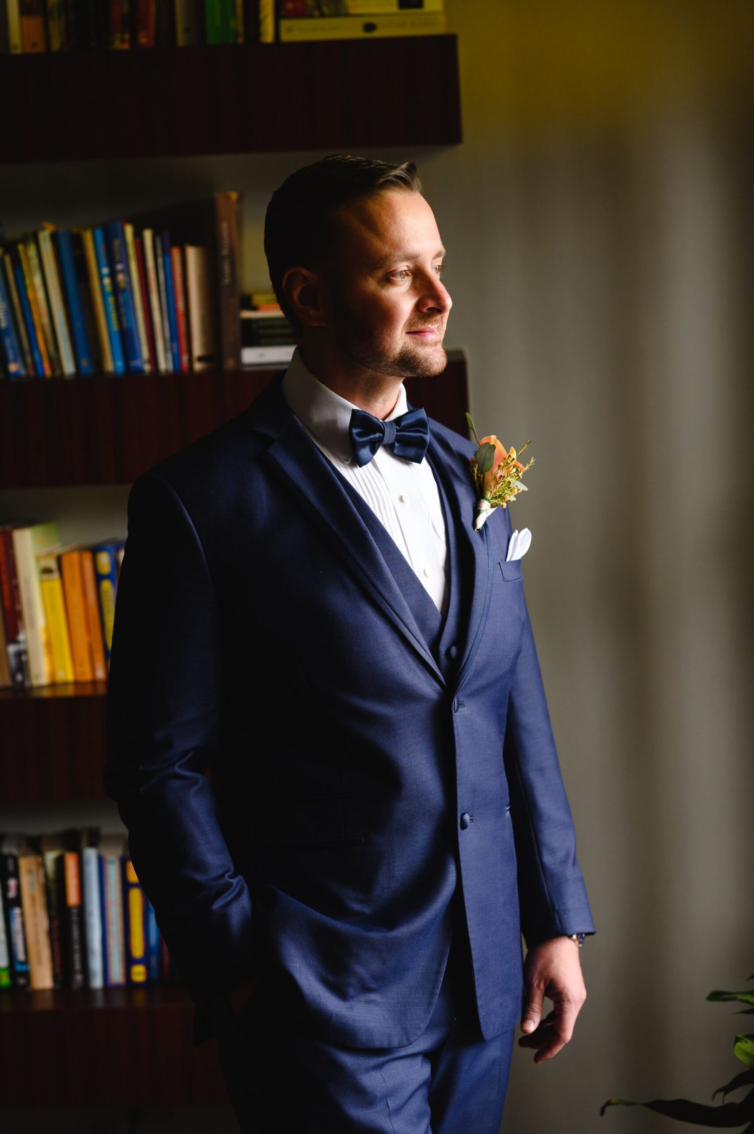 A groom in a navy blue suit with bow tie and boutonniere stands thoughtfully by a bookshelf in warm light.