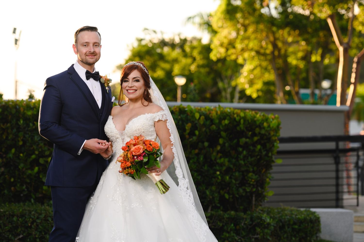 Bride and groom posing together outdoors, she holds orange bouquet in lace gown, he wears navy tux.