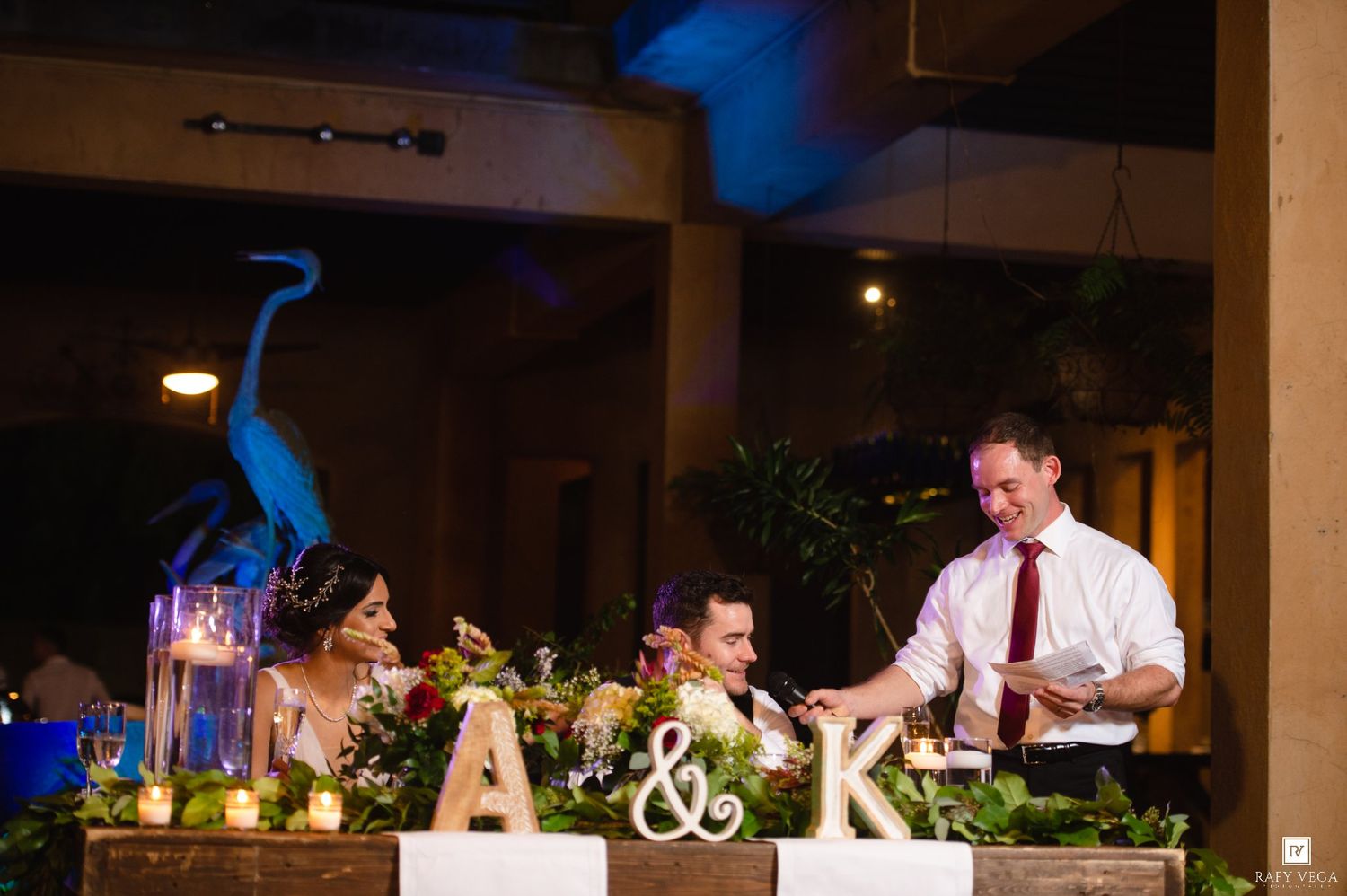 Bride and groom laughing at decorated sweetheart table with floral centerpiece and monogram letters A and K at wedding reception.