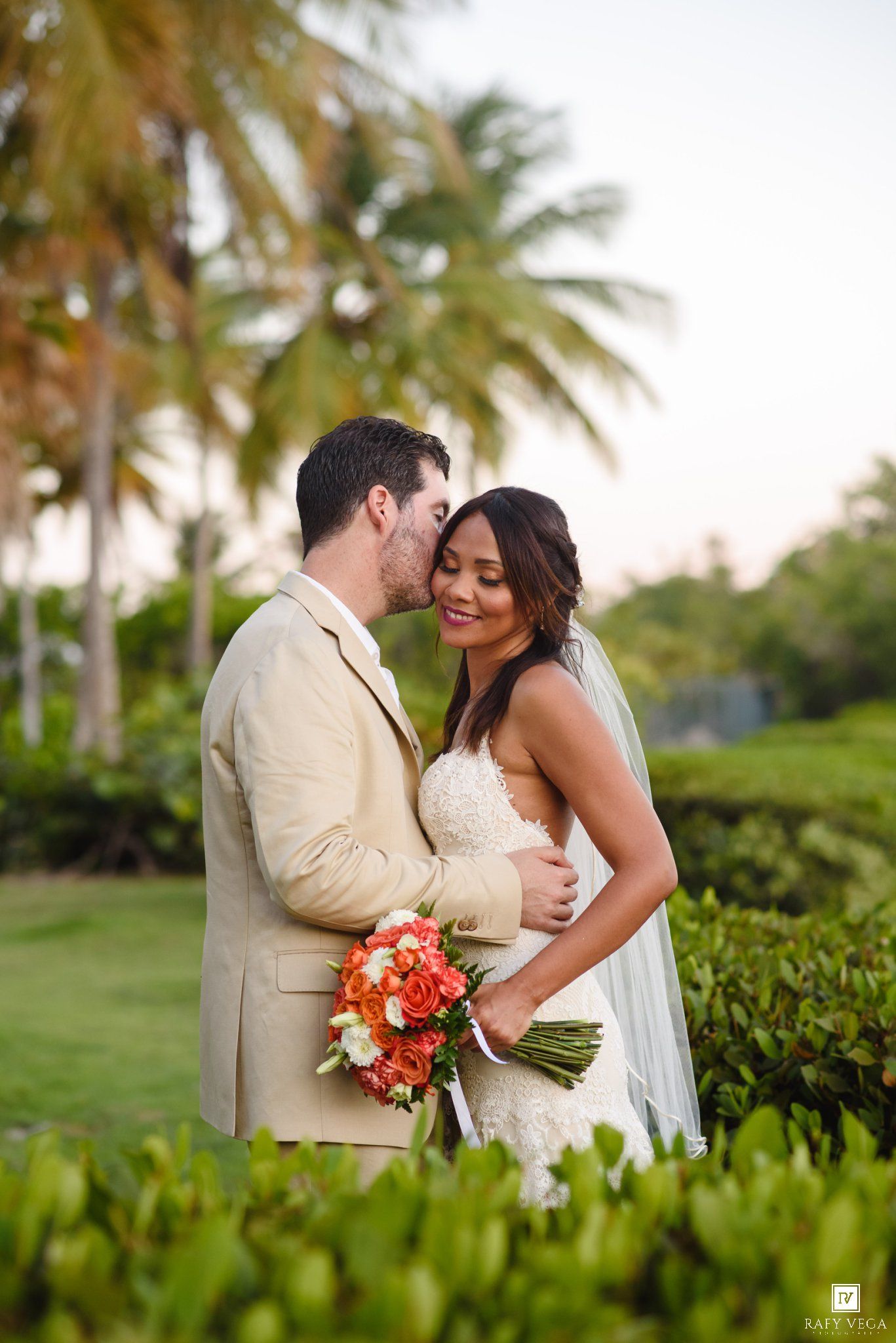 Groom kissing bride on forehead among palm trees, bride holding vibrant orange and red bouquet.