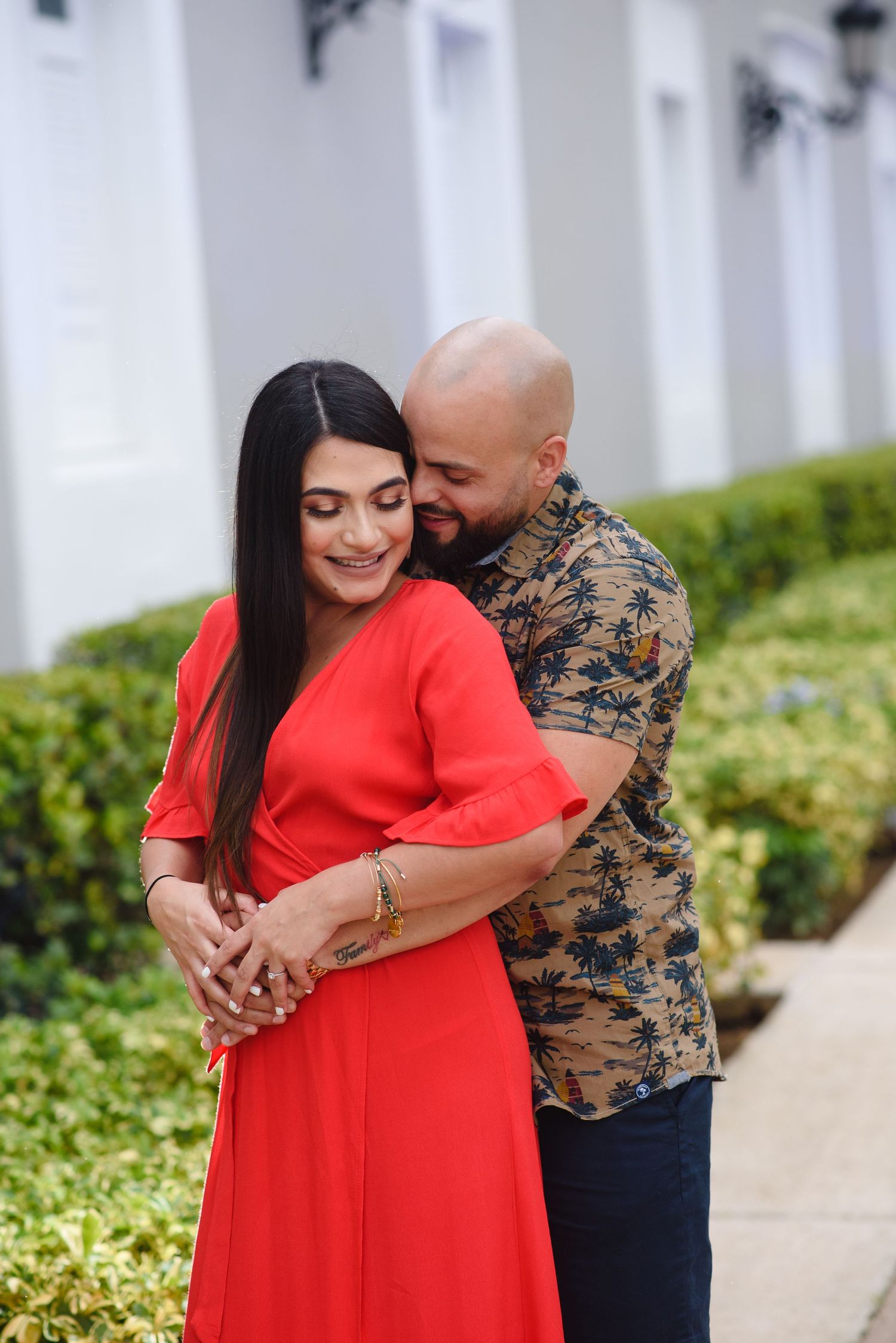 A couple embraces outdoors, woman in red dress, bald man in floral shirt, green hedges and white building behind them.