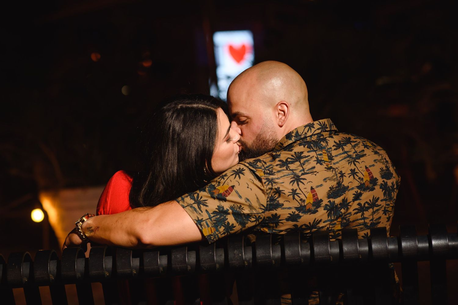 Couple sharing a romantic kiss on a bench at night, man in floral shirt embracing woman in red dress.