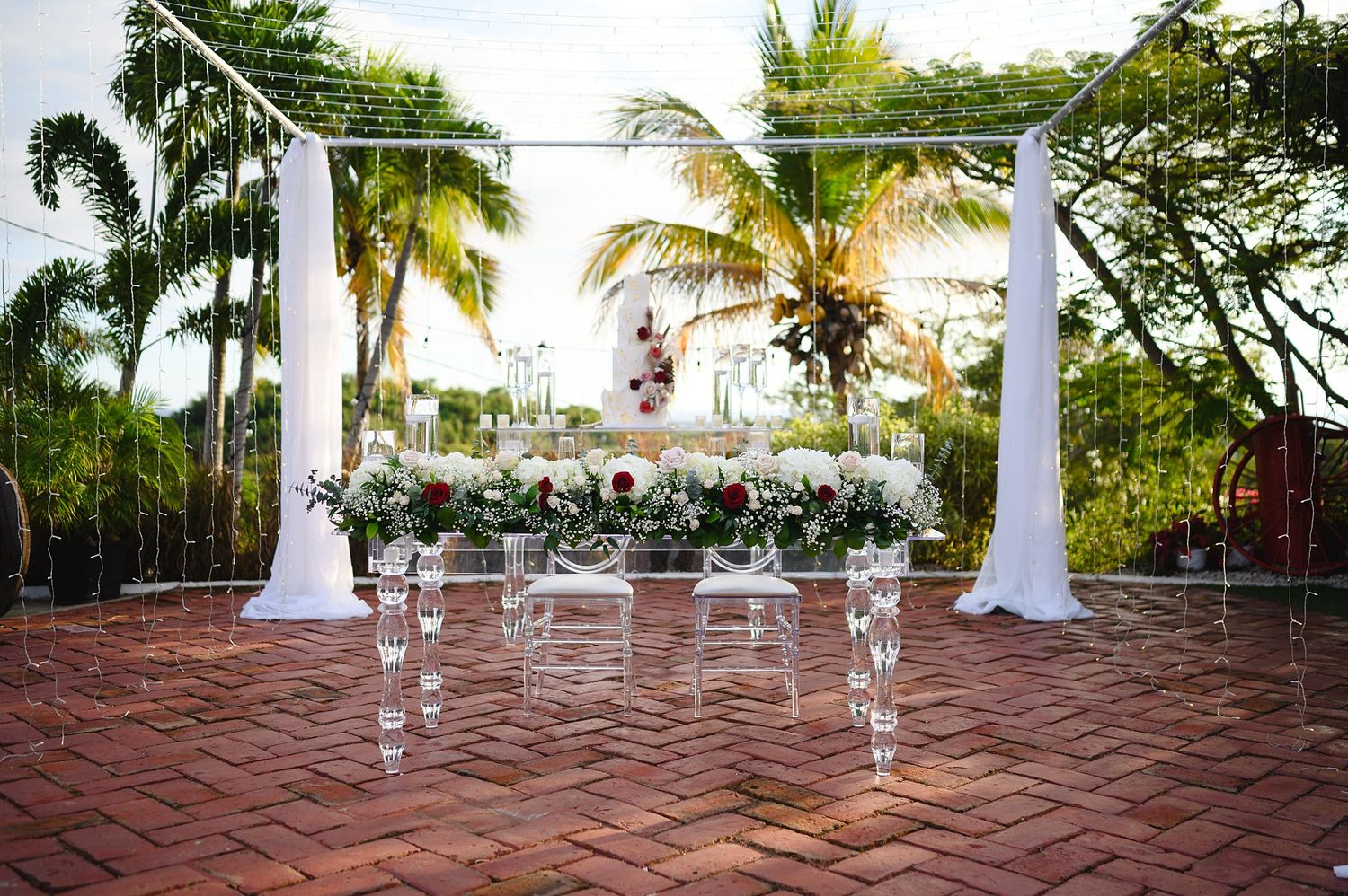 Elegant outdoor wedding altar with white draping, acrylic chairs, and floral arrangements on a brick patio surrounded by palm trees.