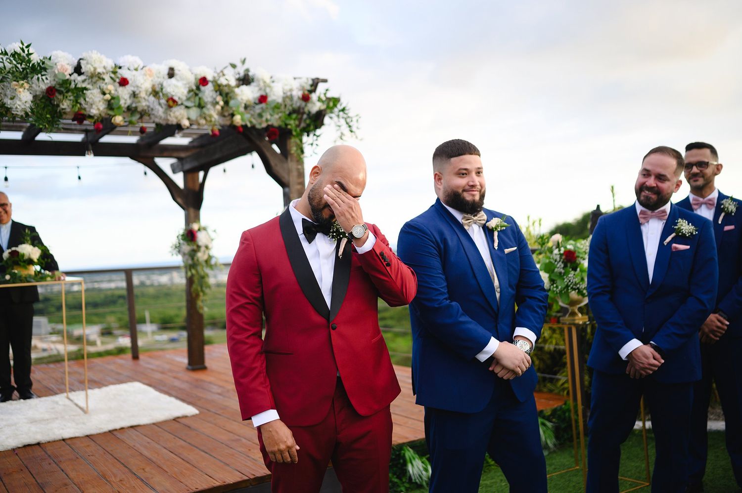 Emotional groom in red suit wiping tears at outdoor wedding ceremony with groomsmen in navy blue suits.