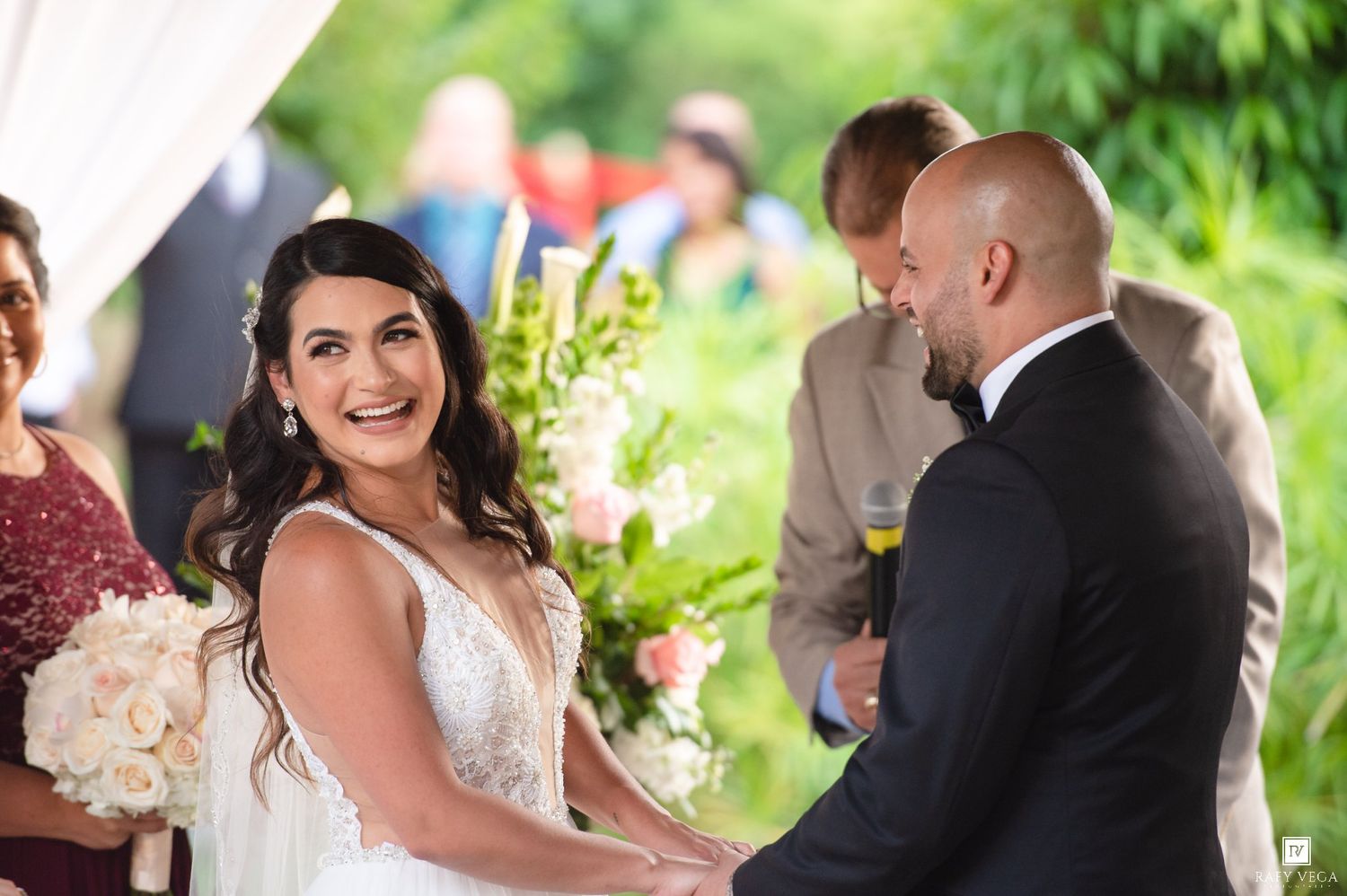 Bride smiling joyfully at groom during outdoor wedding ceremony with floral arrangements.