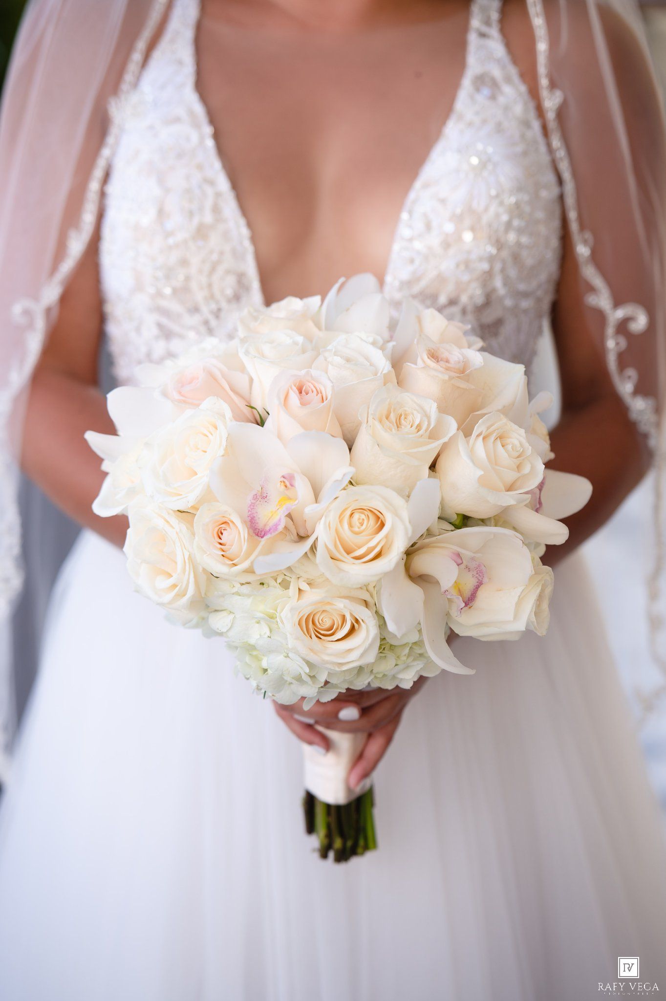 Bride holding elegant white rose and orchid wedding bouquet, wearing beaded lace gown with deep V-neckline and veil.