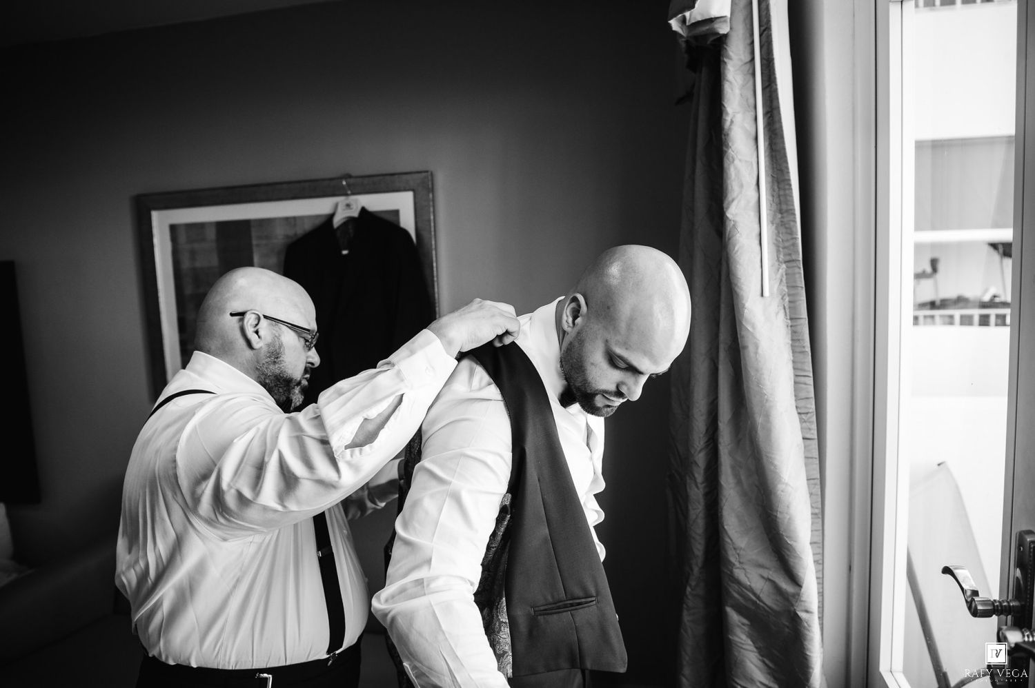 Black and white photo of two bald men in suits, one adjusting the other's bow tie before a wedding ceremony.