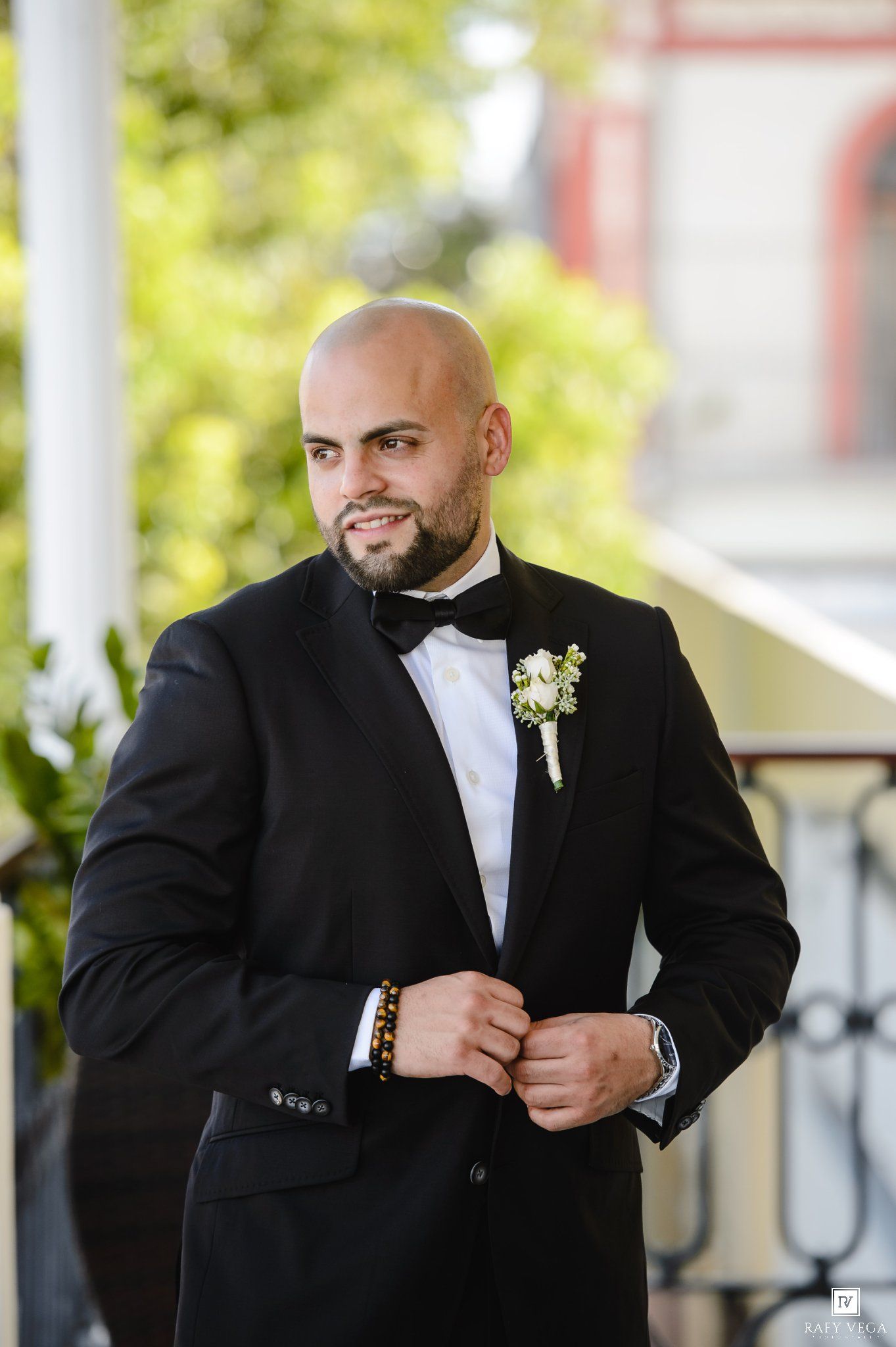 Bald groom in black tuxedo with bow tie and boutonniere adjusting jacket on outdoor balcony.