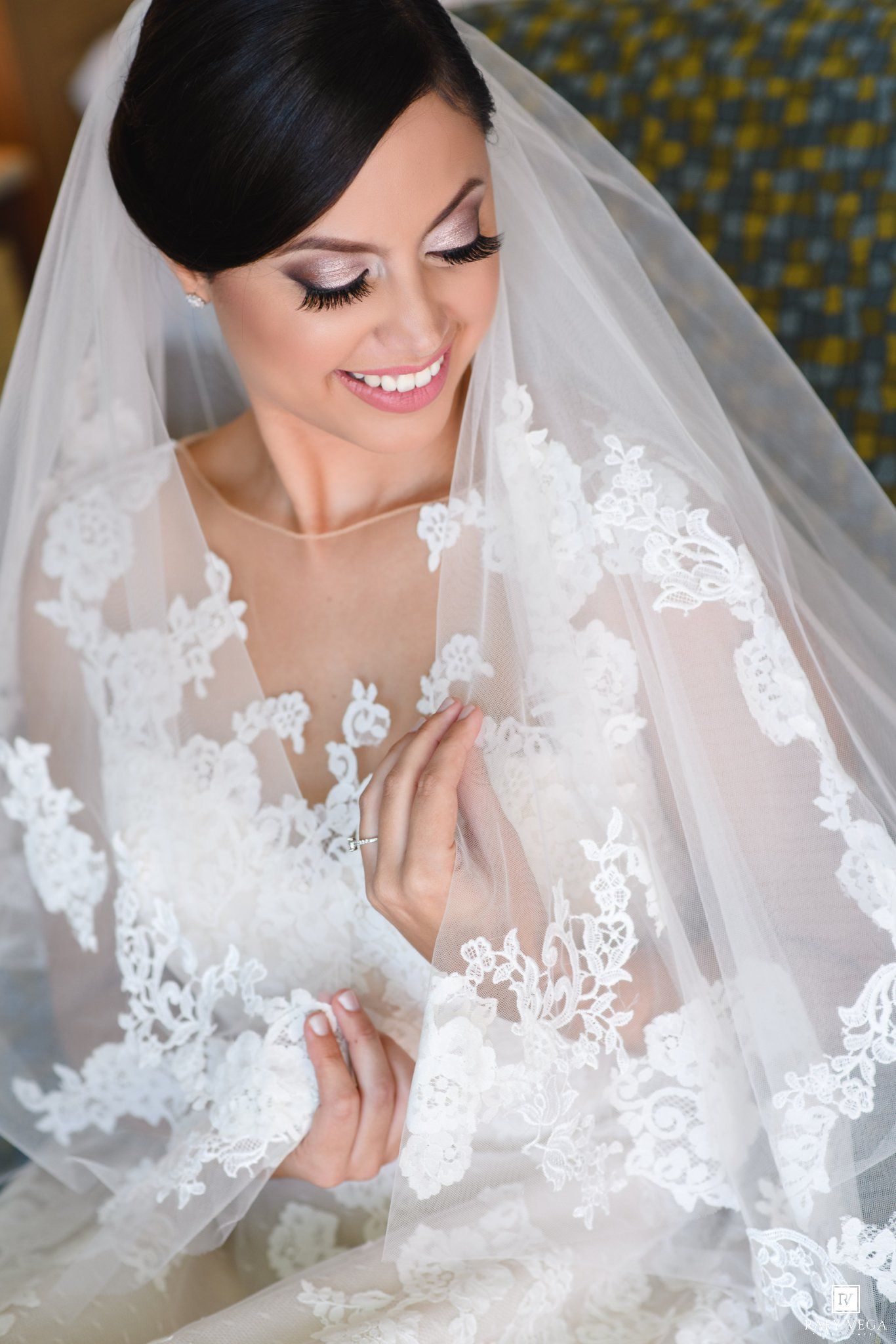 Smiling bride in lace wedding dress and veil, with elegant makeup and dark hair, looking down gracefully.