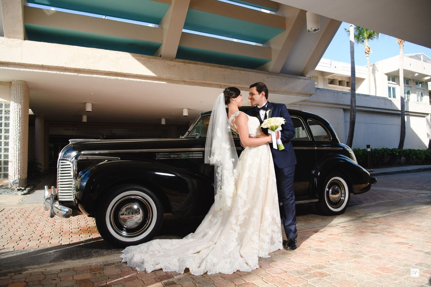 Bride and groom share a kiss beside a vintage black car on their wedding day.