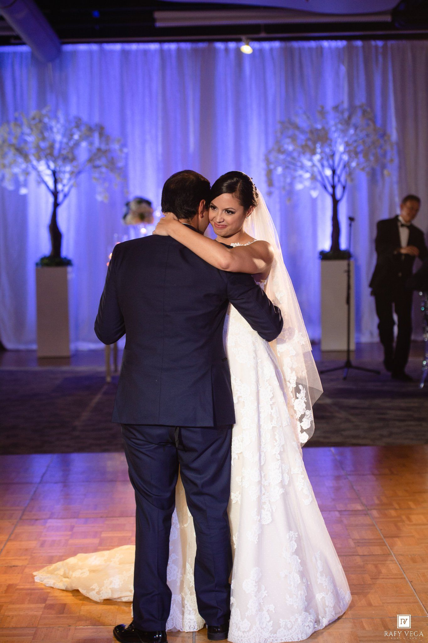 Bride and groom share their first dance at elegant wedding reception with white drape backdrop and decorative trees.