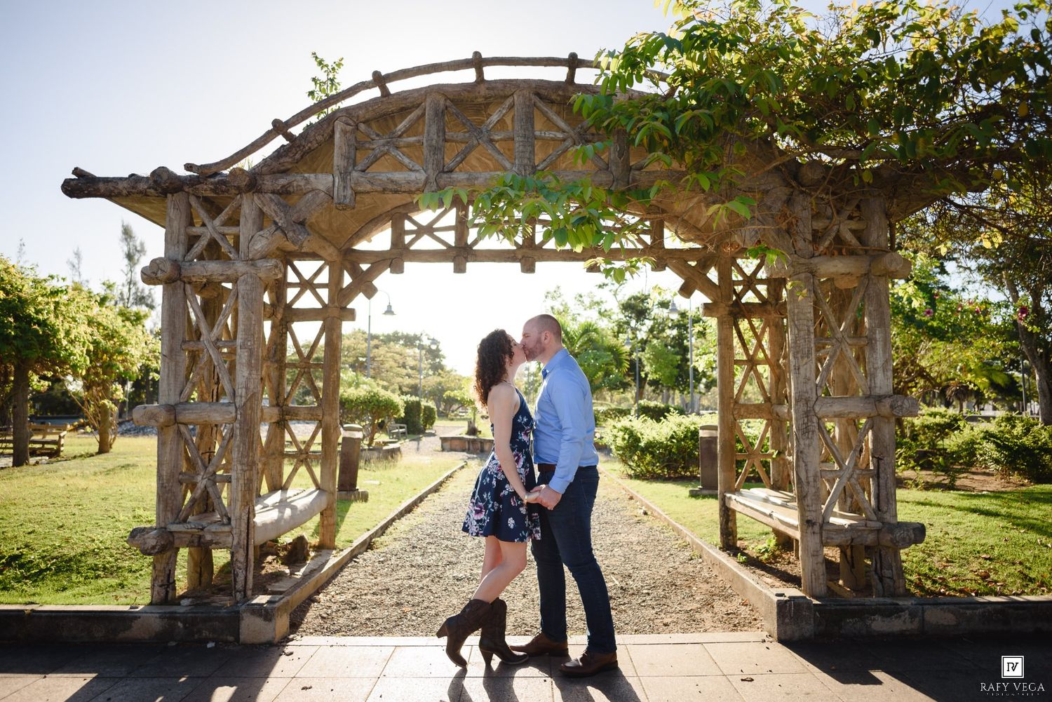 Couple sharing a romantic moment under a rustic wooden garden arch surrounded by lush greenery on a sunny day.