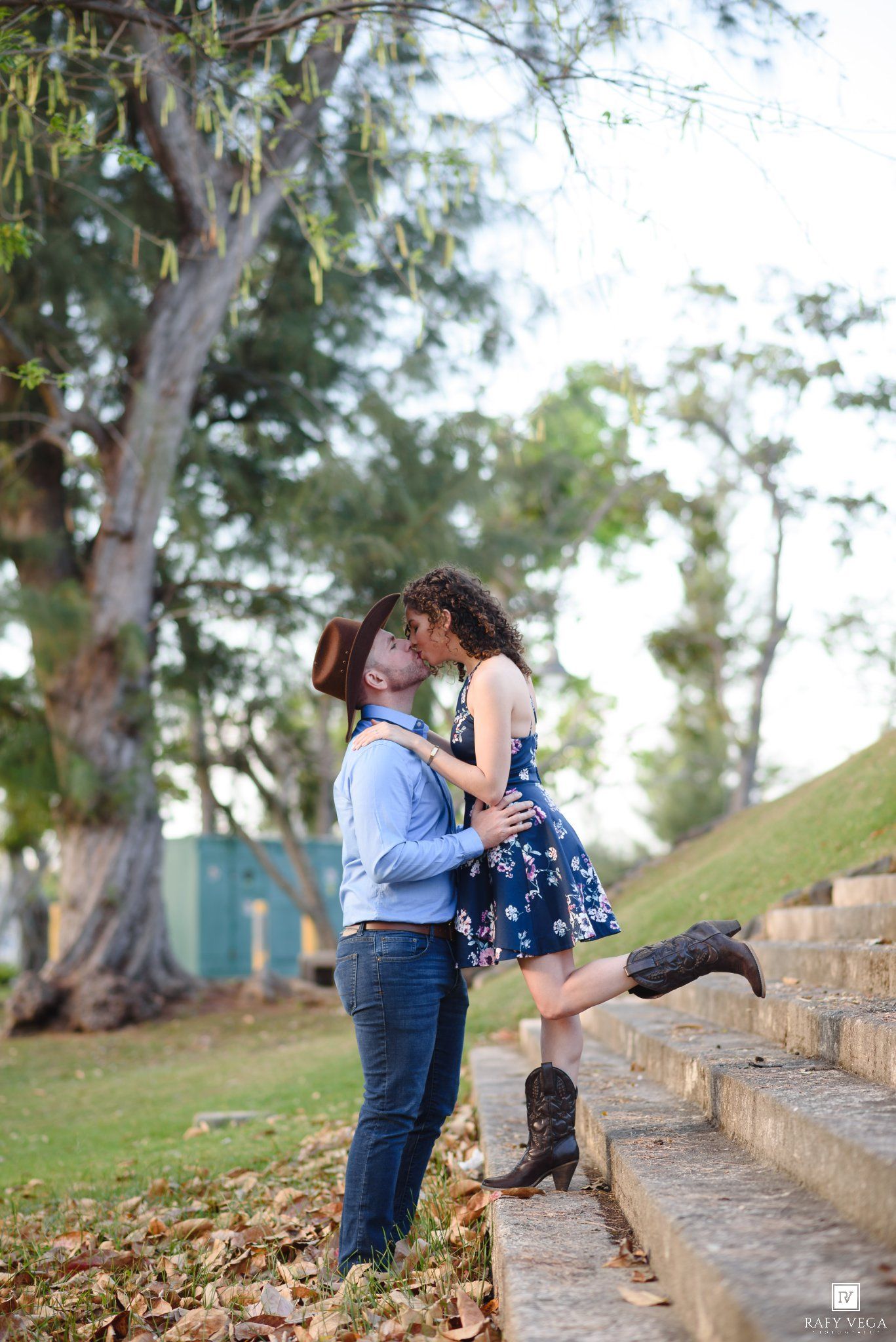 Couple sharing a romantic kiss on stone steps outdoors, woman in floral dress and cowboy boots, man in blue shirt and hat.