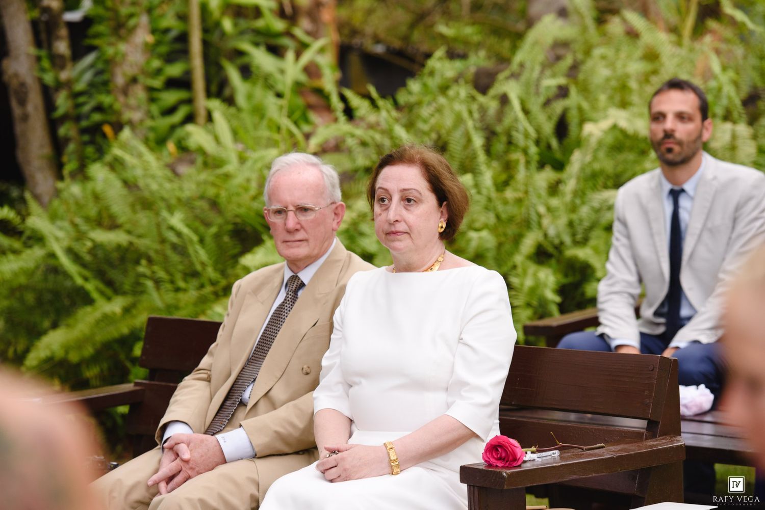 Elegant couple seated at outdoor wedding ceremony with ferns in background, man in tan suit, woman in white.