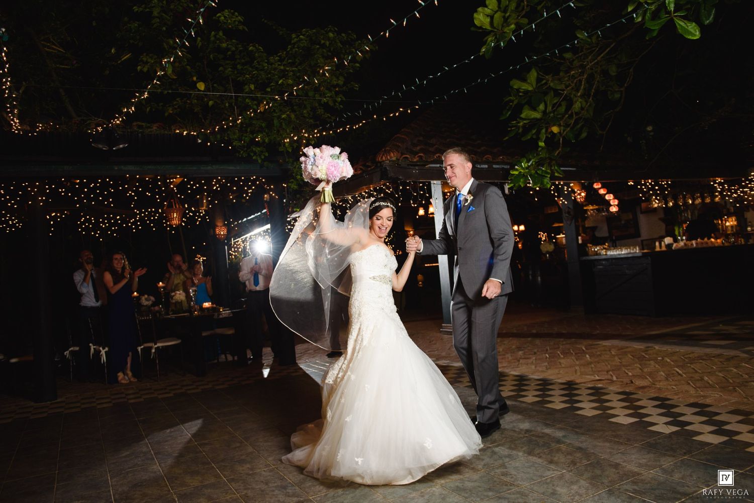 Bride and groom make grand entrance at outdoor evening wedding reception with string lights and checkered dance floor.