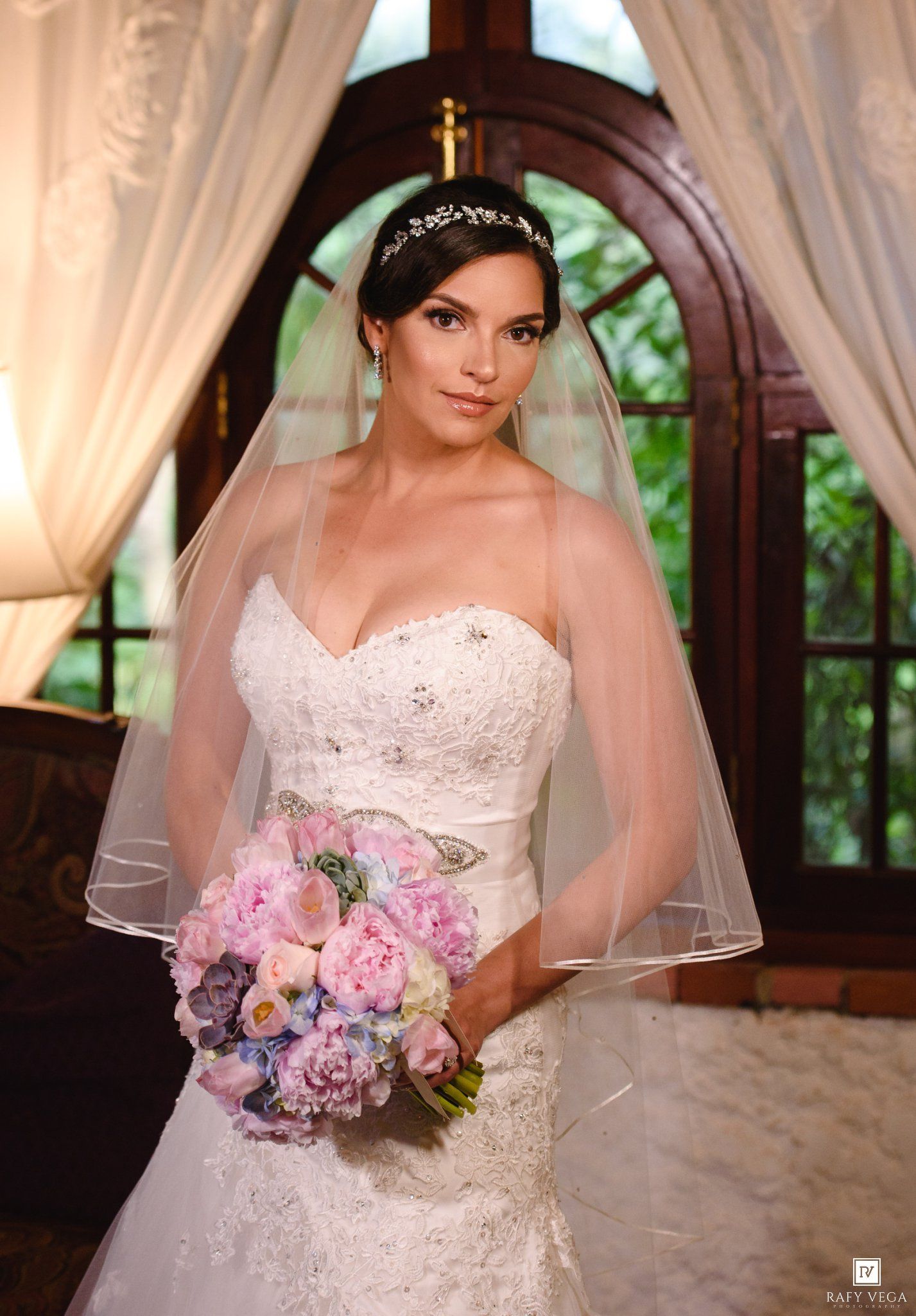 Elegant bride in strapless lace gown with veil and tiara holding pink and lavender bouquet indoors.