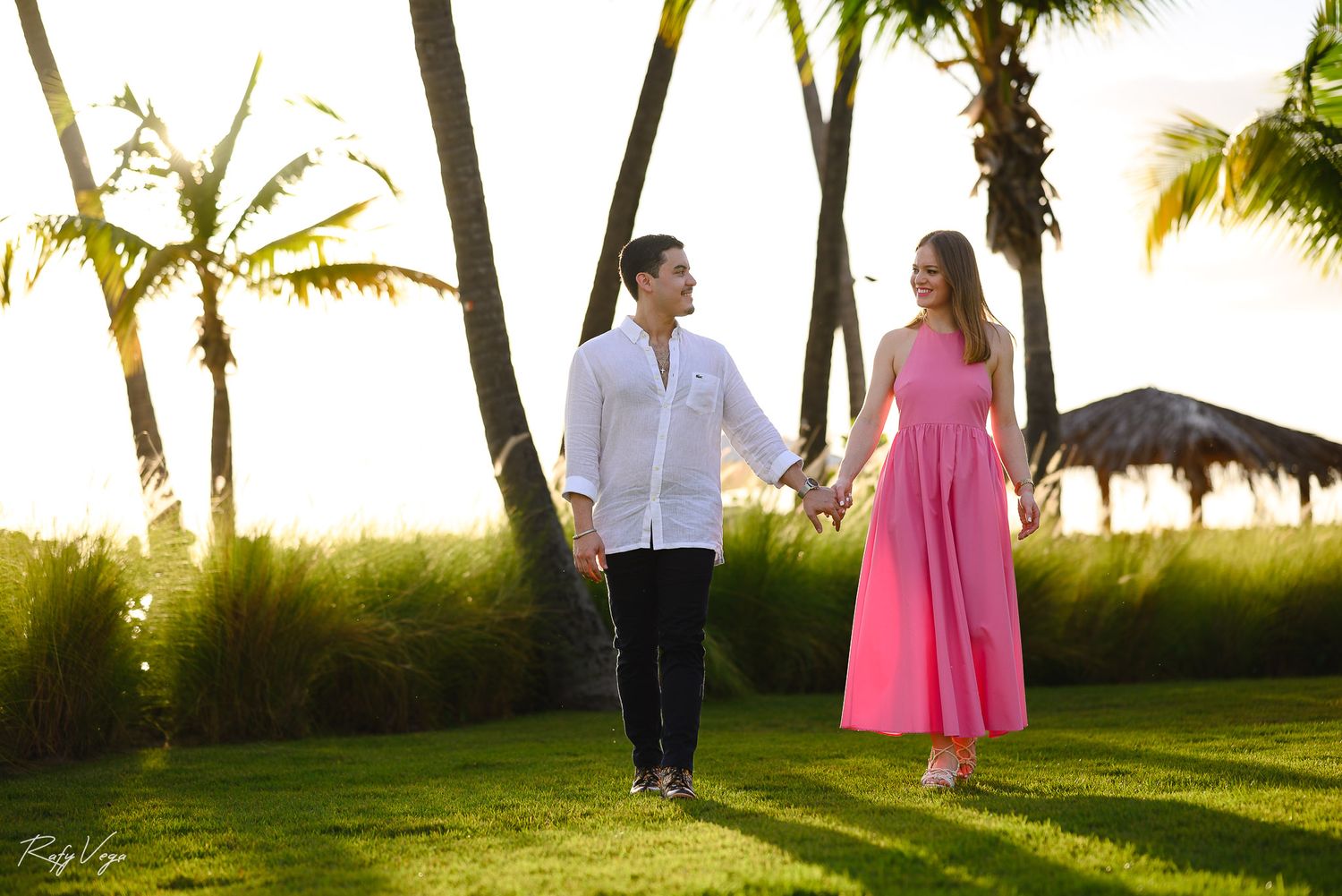Couple holding hands walking on tropical resort lawn at sunset, woman in pink dress, man in white shirt, palm trees background.