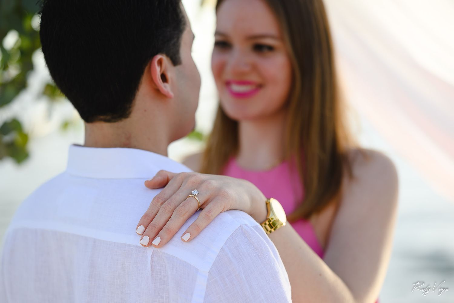 Smiling woman in pink dress places hand with engagement ring on man's shoulder during romantic outdoor moment.