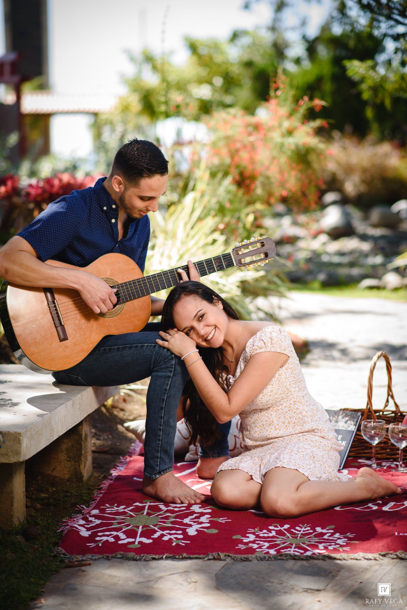 A man serenades a woman with acoustic guitar during a romantic outdoor picnic on a red blanket.